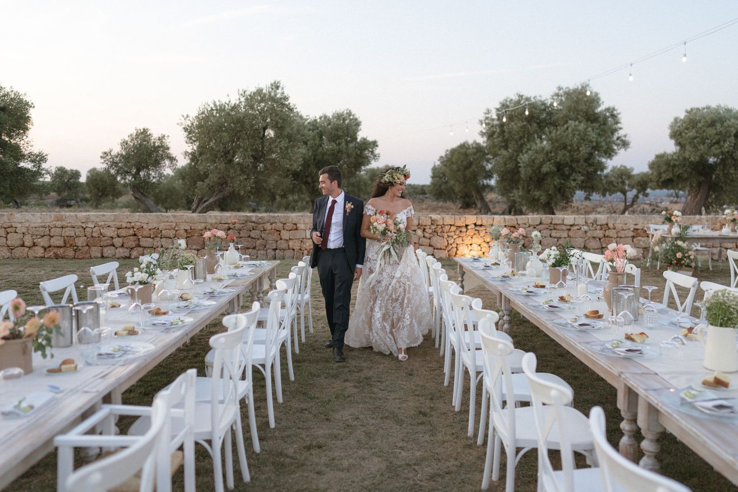 Elegant outdoor wedding reception setup with long tables and white chairs in an olive grove.