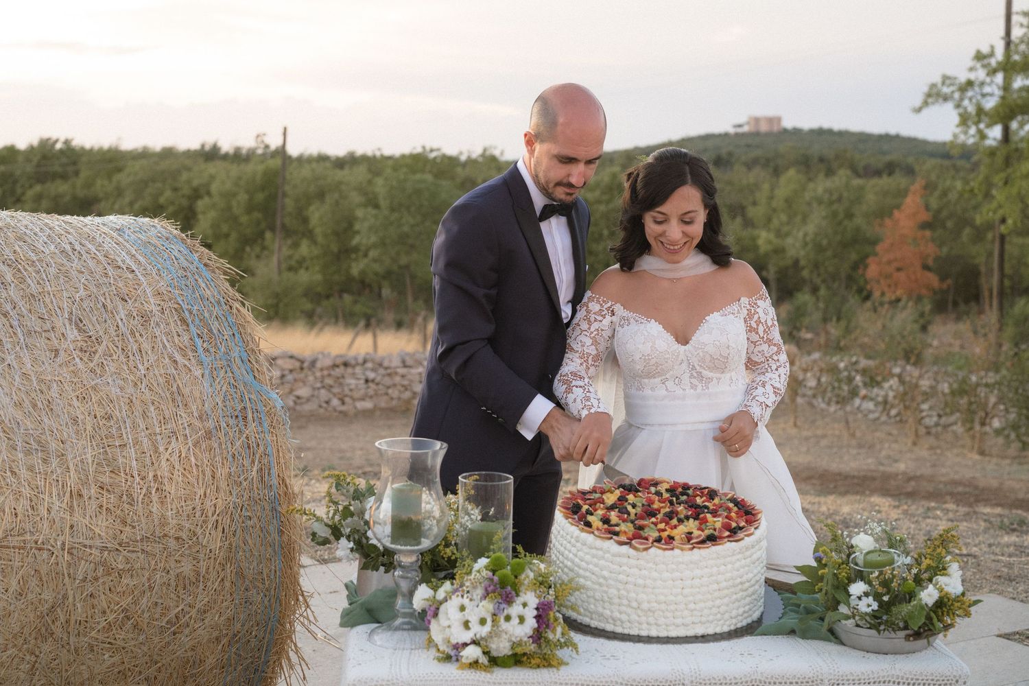 A rustic outdoor wedding reception setup with hay bales, string lights, and floral decorations at dusk.