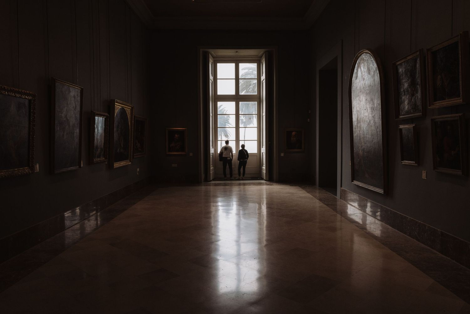 Dark museum hallway with sunlit window at the end and artwork hanging on walls.