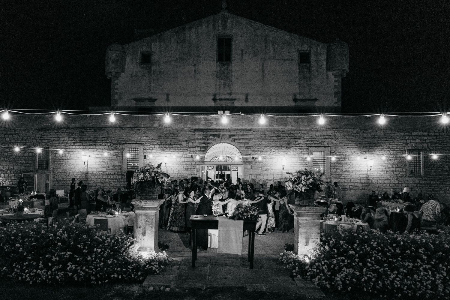 An outdoor evening wedding reception with string lights illuminating a historic stone building.