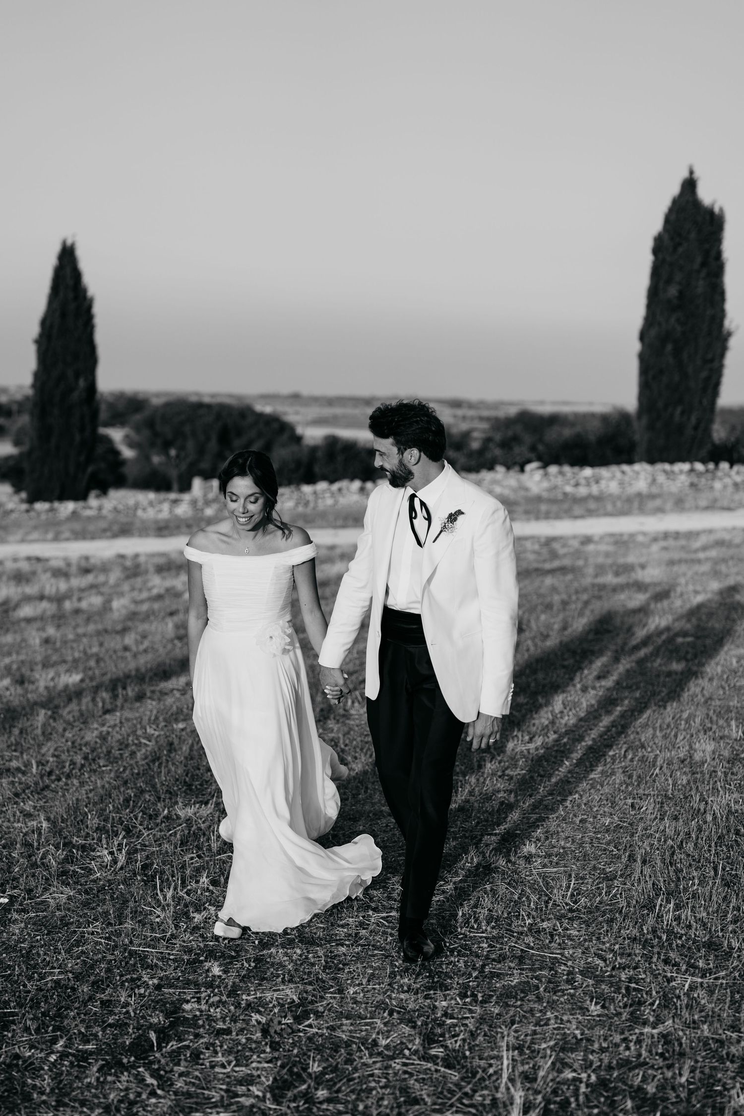 A couple in wedding attire walks through a scenic countryside field with cypress trees in the background.