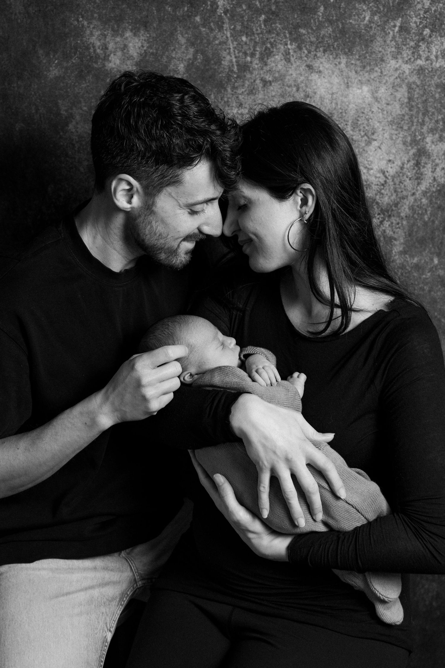 Black and white photo of couple touching foreheads while holding their newborn baby in a studio portrait.