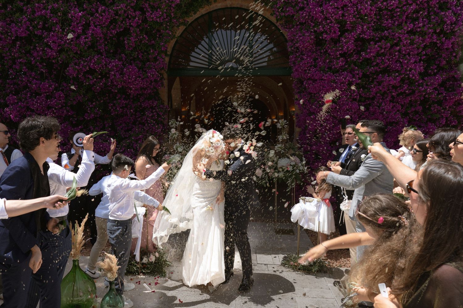 Bride and groom share a kiss as guests celebrate with confetti outside a venue covered in purple bougainvillea.
