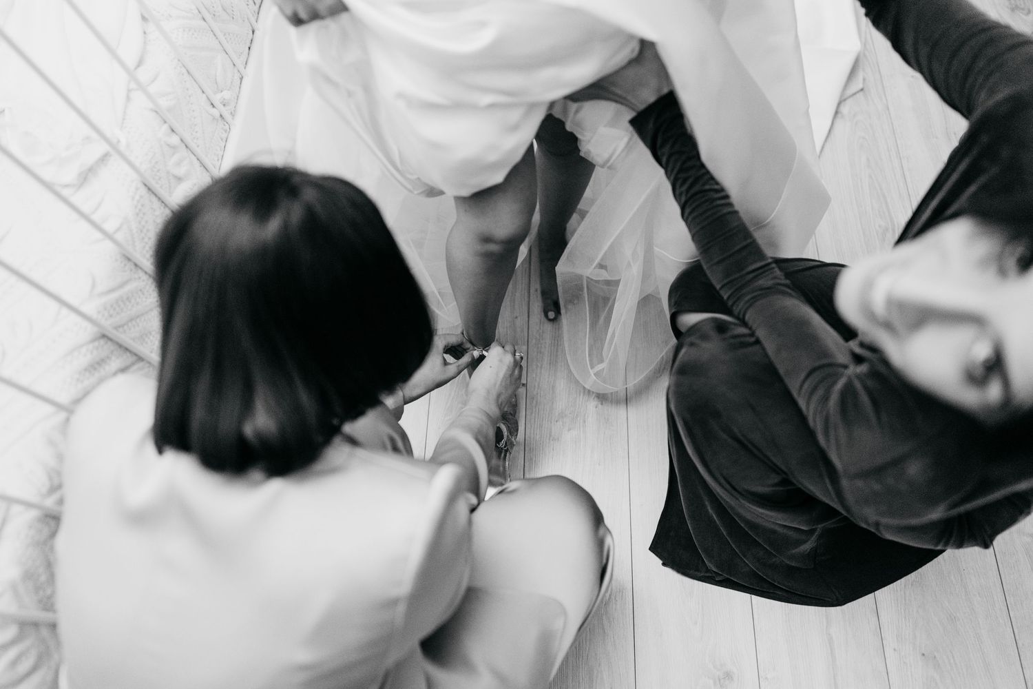 Overhead black and white shot of bridesmaids helping a bride with her wedding dress and shoes on the floor.