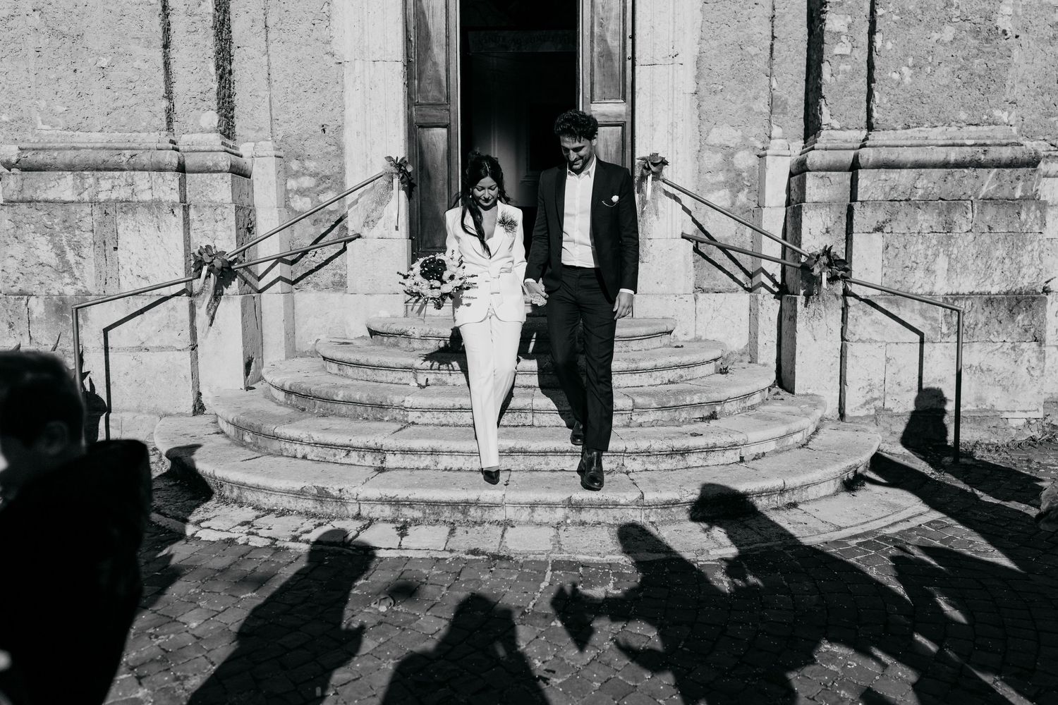 Bride in white and groom in dark suit walk down historic stone church steps, their long shadows cast across the sunlit pavement.