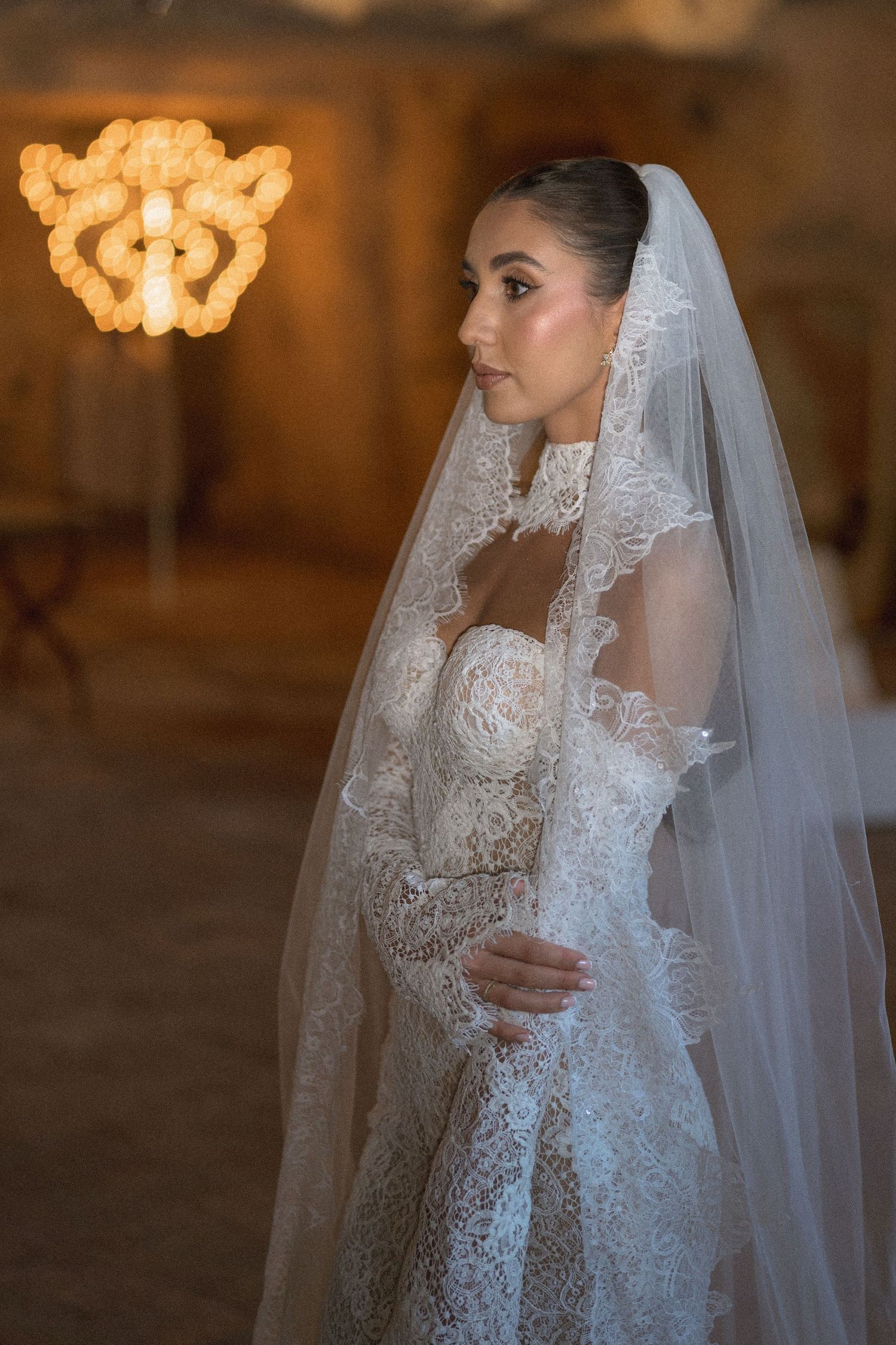 Bride in a lace wedding gown and lace-edged cathedral veil standing in a warmly lit room with a glowing chandelier.