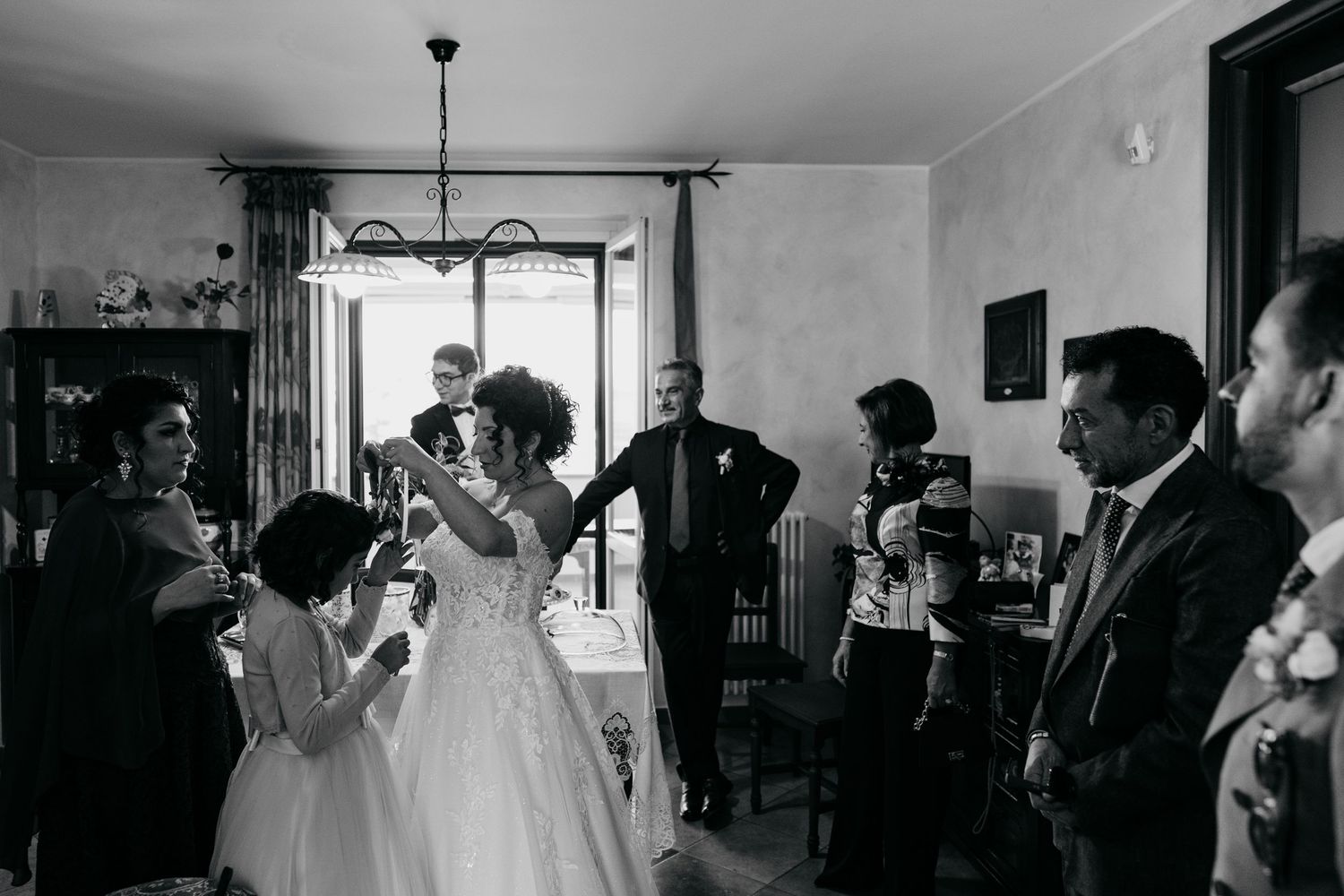 Black and white wedding photo of bride and groom toasting with guests in elegant home interior.