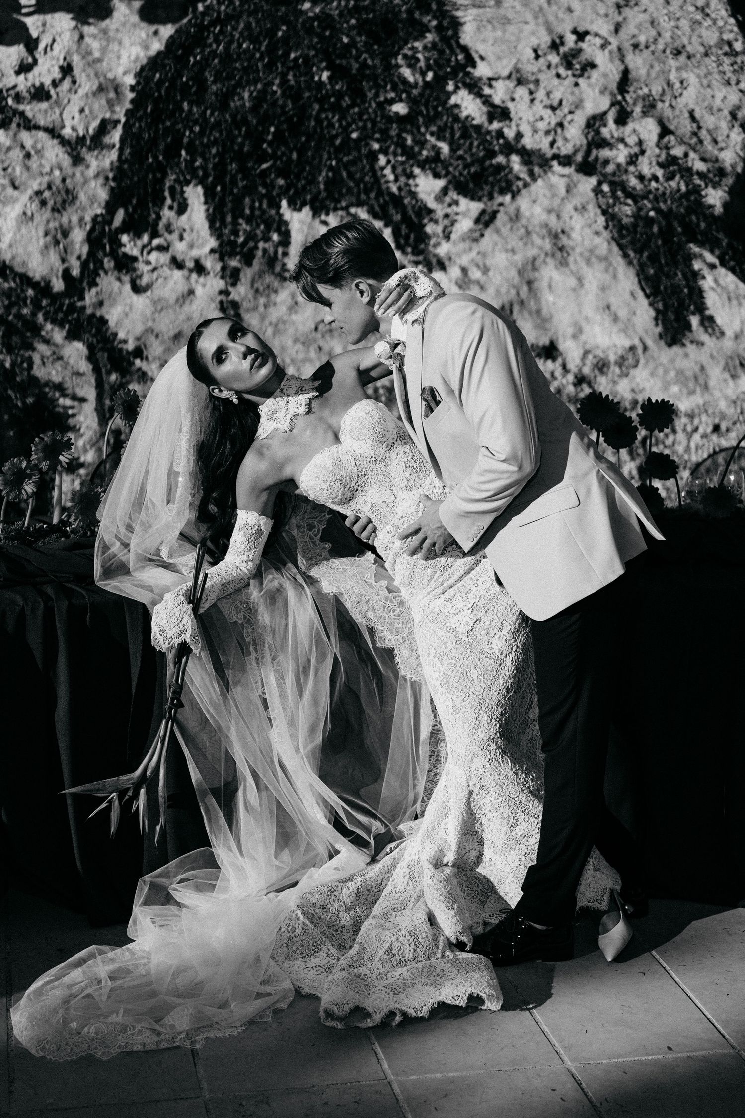 Black and white image of a groom dipping his bride in a lace wedding dress and veil against a rocky, ivy-covered wall.