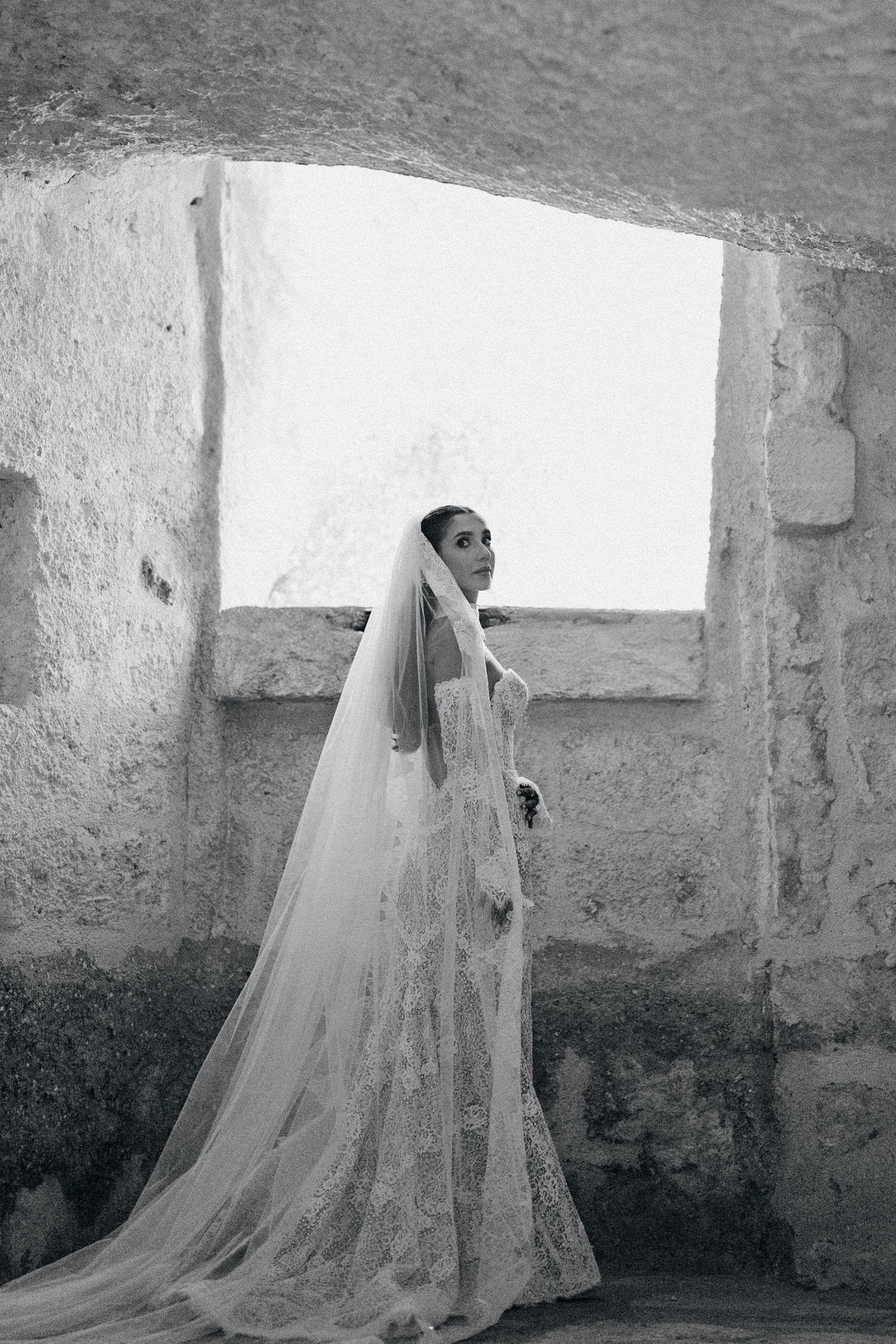 Black and white portrait of a bride in a lace wedding gown and long veil standing by a bright window in an ancient stone cave.