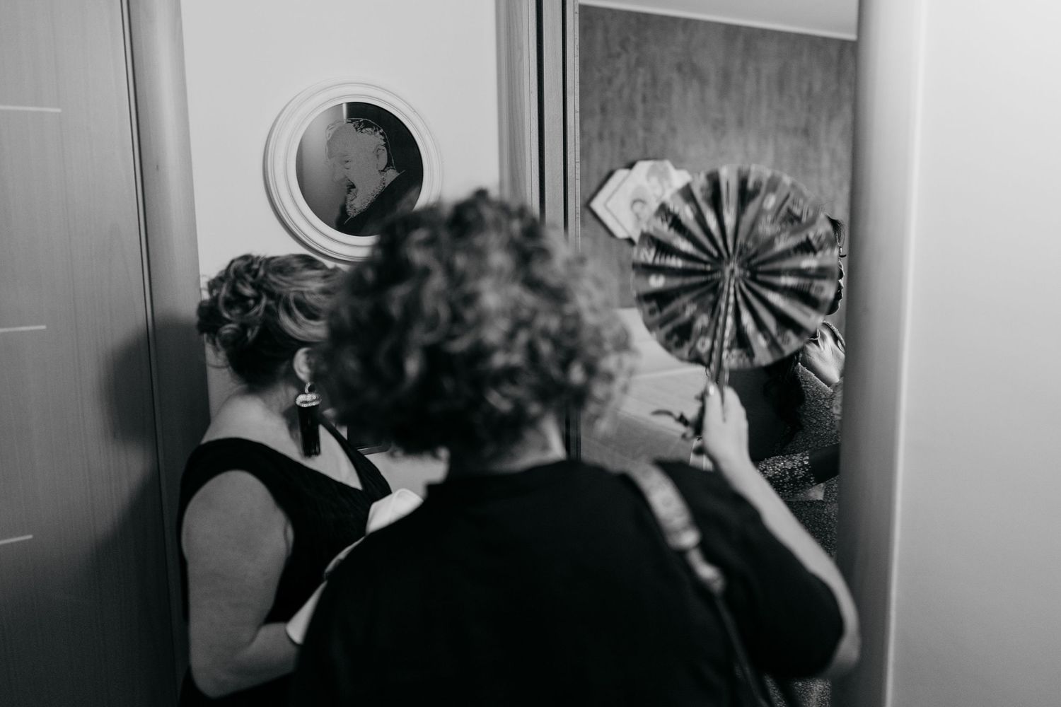 Black and white photo of two women, one holding a decorative fan, preparing in front of a mirror.
