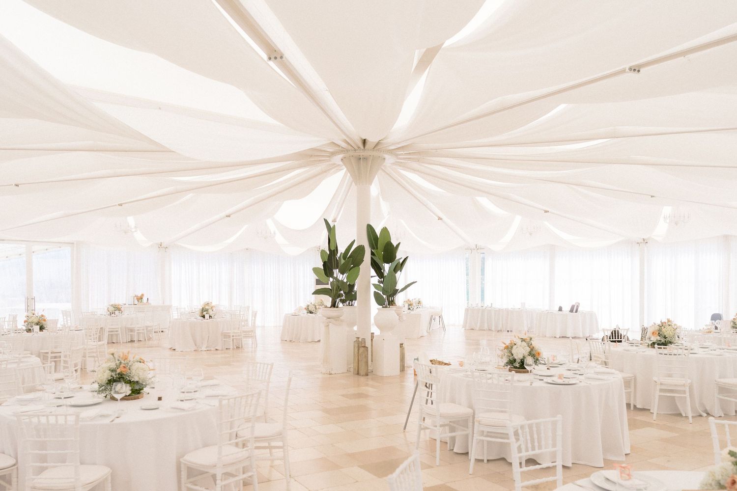 Elegant all-white wedding reception venue with round tables under draped ceiling.