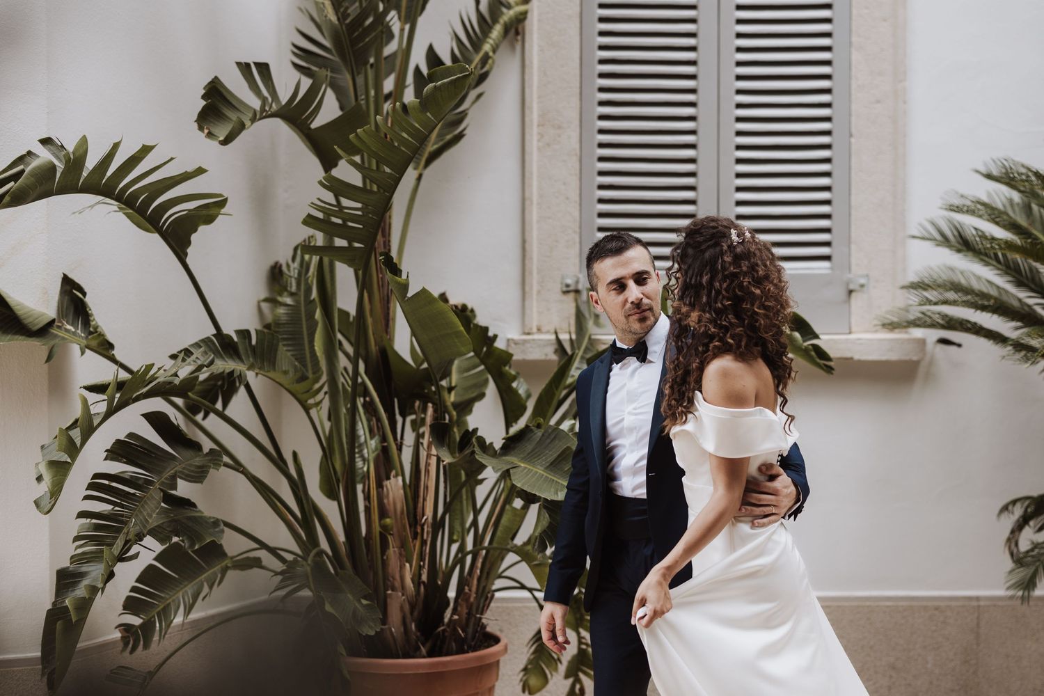 A romantic wedding moment against a white wall with tropical palm plants framing the elegant scene.