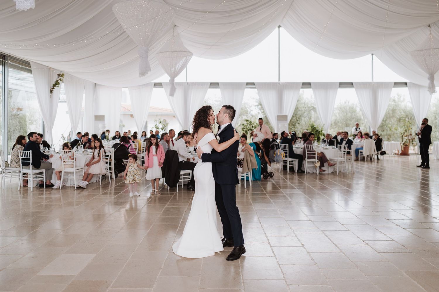 A couple shares their first dance in an elegant white tent reception space surrounded by guests.