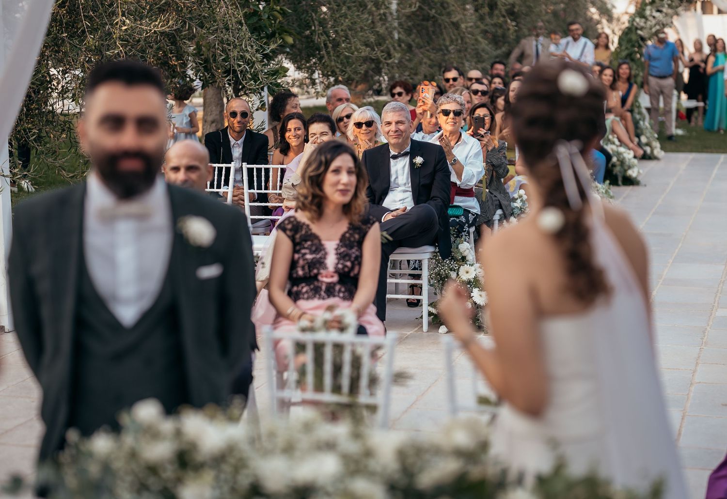 Wedding guests watch ceremony from white chairs lined up outdoors with floral decor.