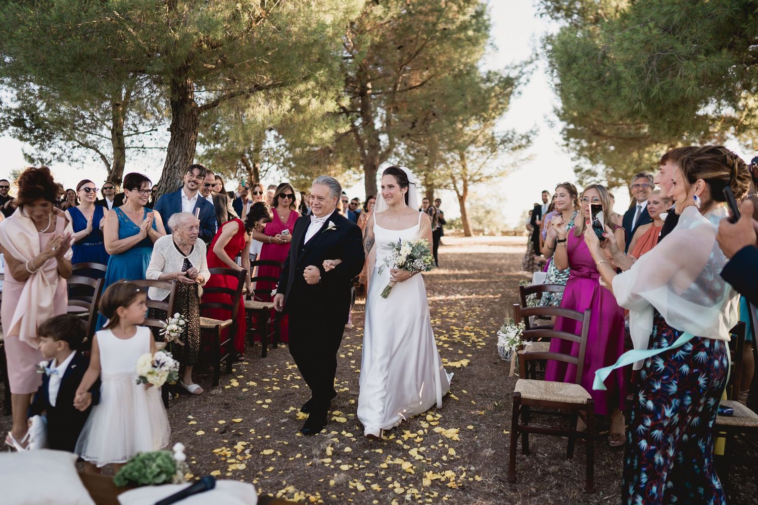 Wedding celebration with guests dressed in colorful attire standing among pine trees during outdoor ceremony.