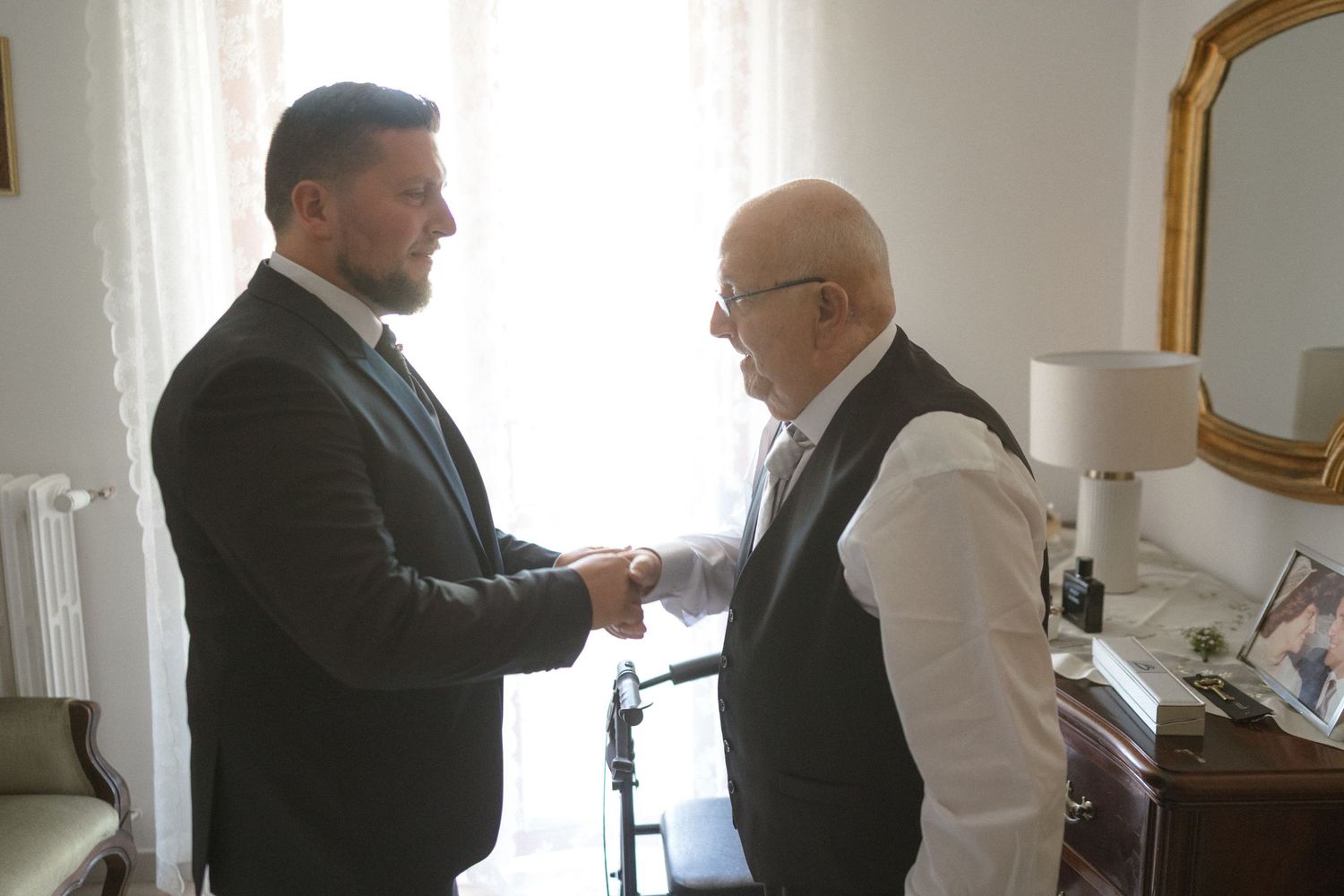 Two men in business suits shake hands in an office setting with white walls and decorative mirrors.