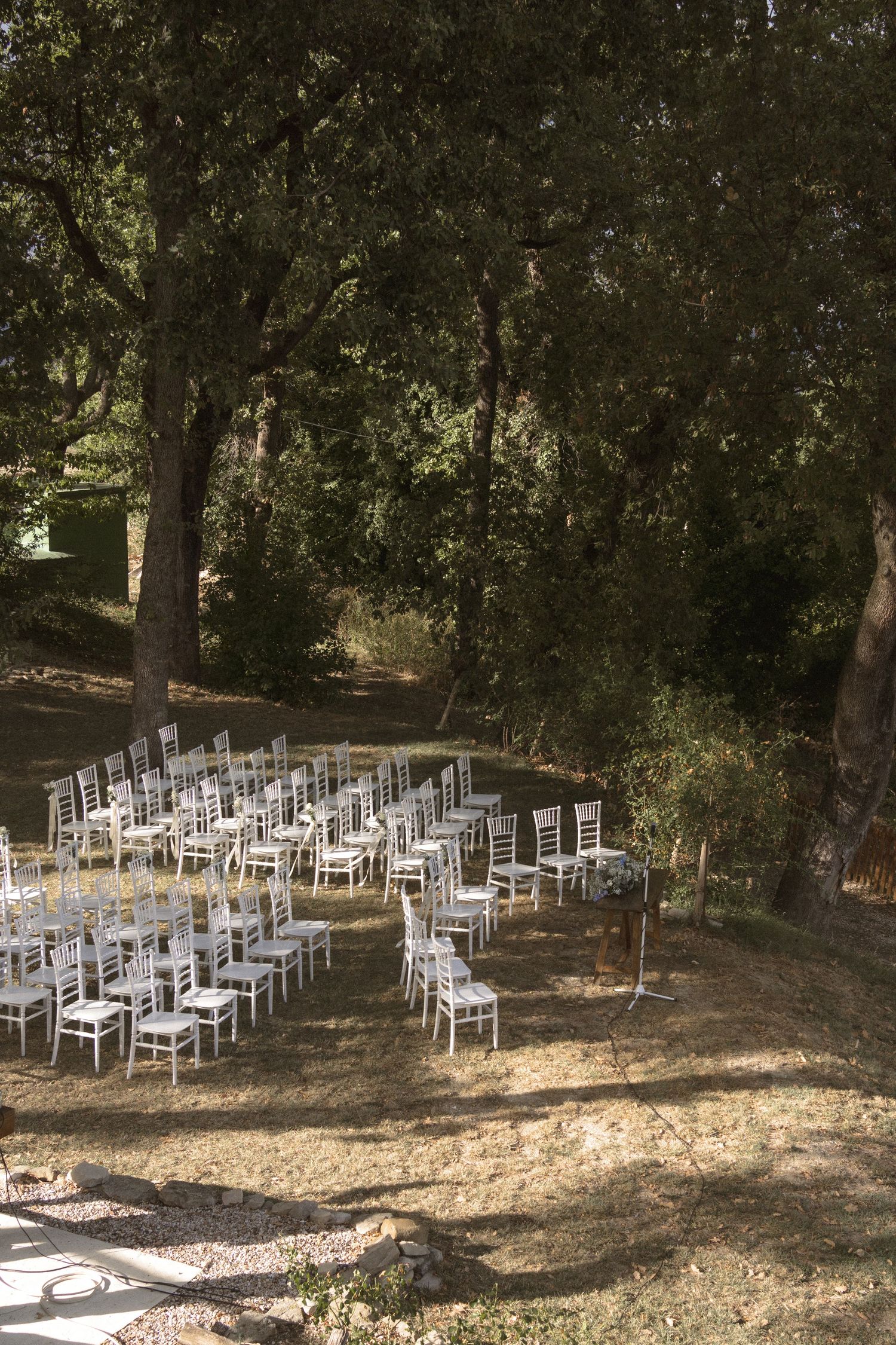 Rows of white chairs arranged in a wooded outdoor setting for a wedding ceremony under shaded trees.
