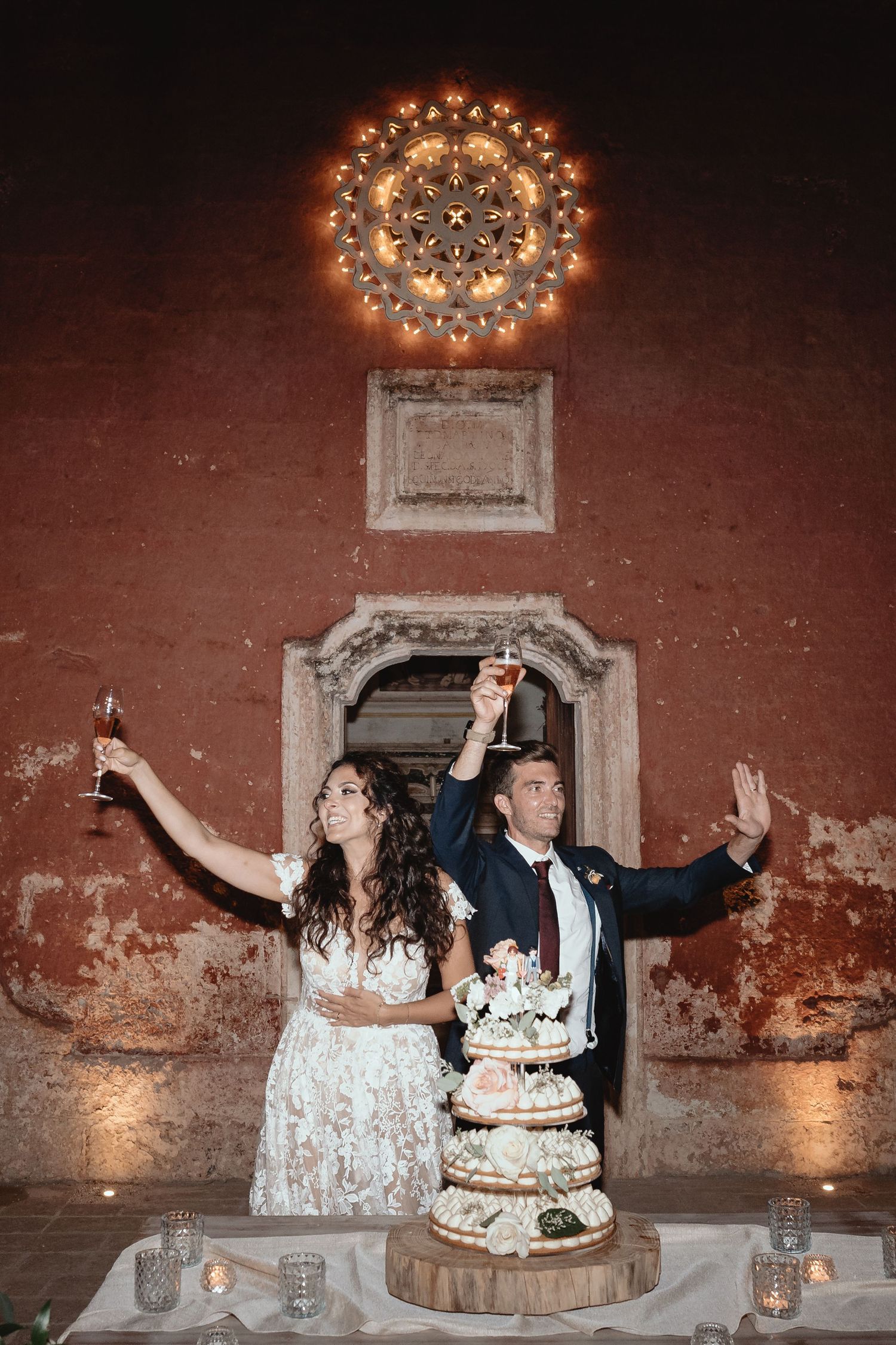 Wedding couple posing with their cake against a rustic brick wall with elegant lighting.