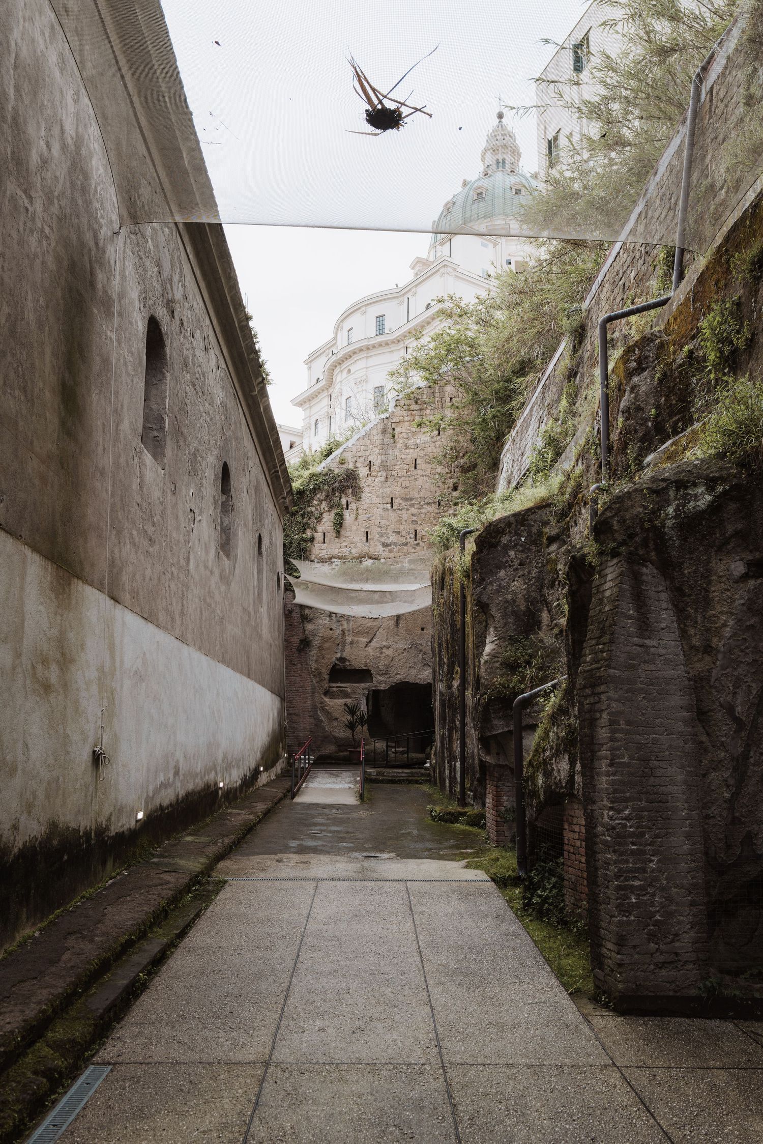 Narrow stone pathway between historic walls leading to a domed building with a helicopter overhead.