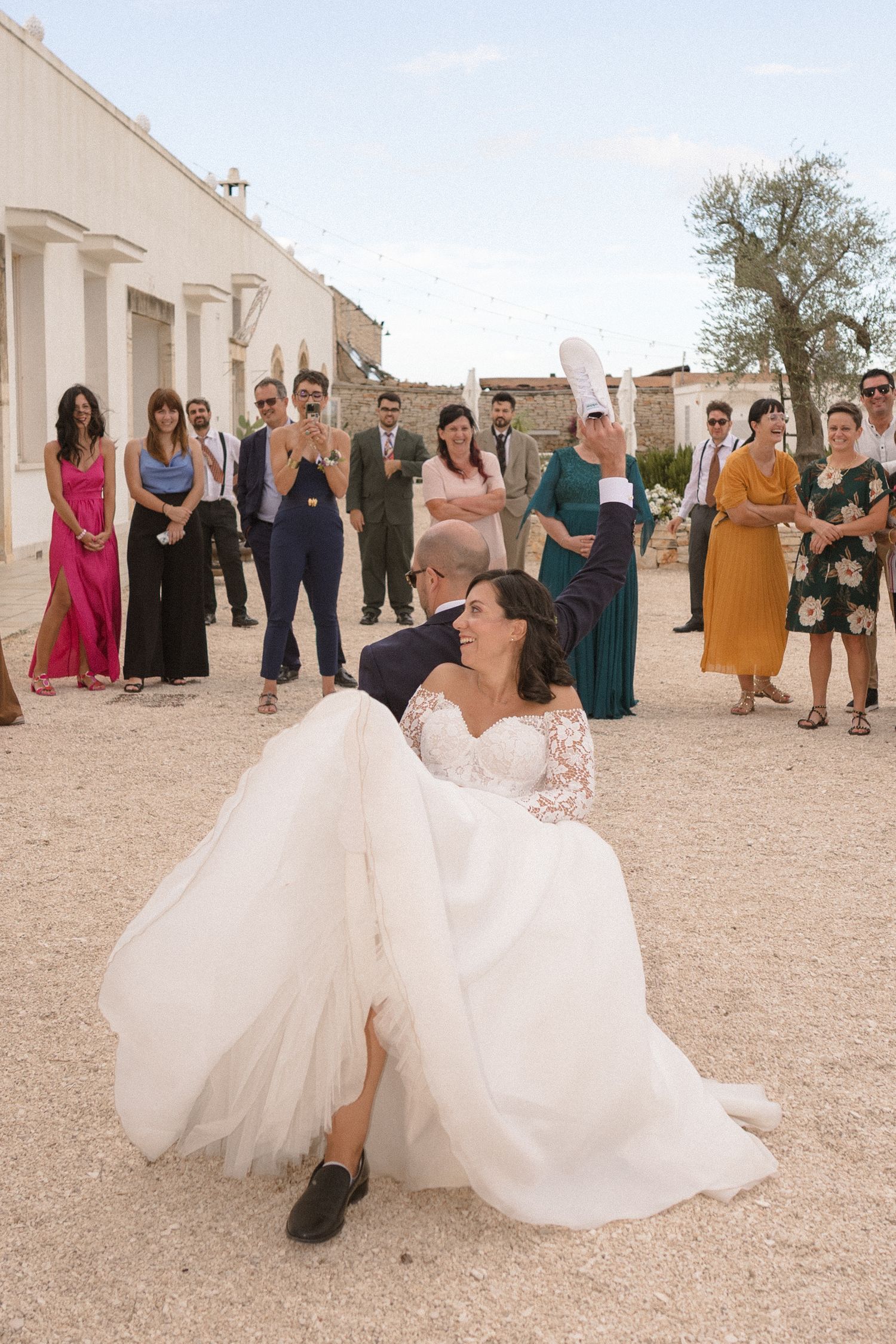 Bride in flowing white dress twirls on desert grounds surrounded by wedding guests.