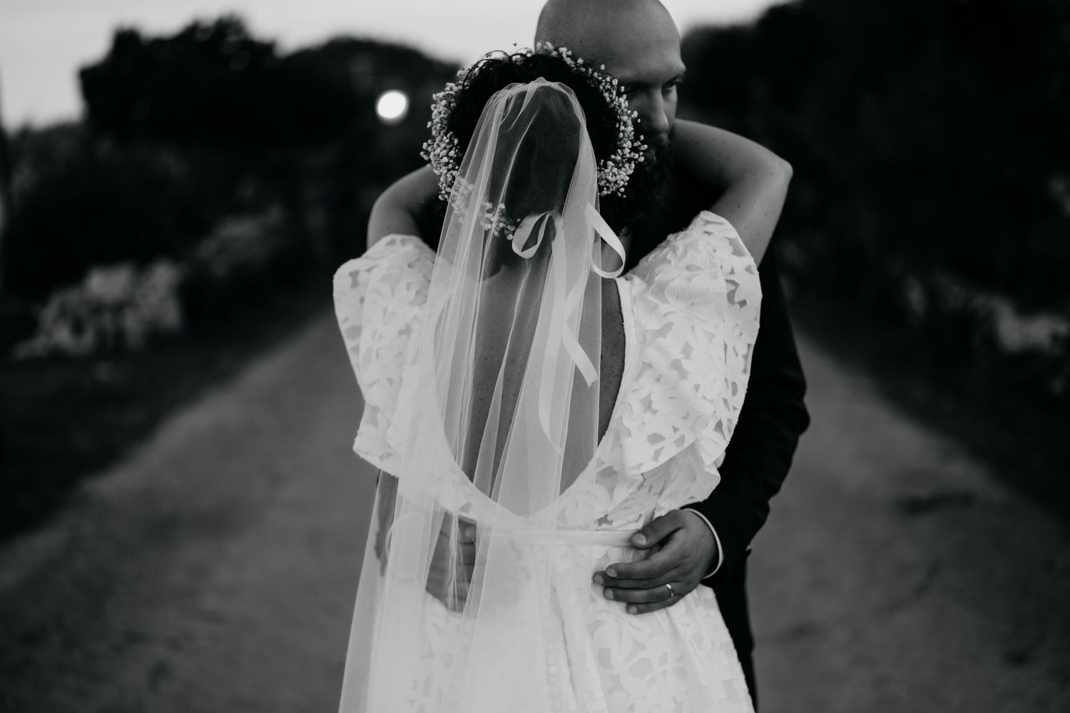 Black and white photo of a couple in wedding attire walking along a tree-lined path.