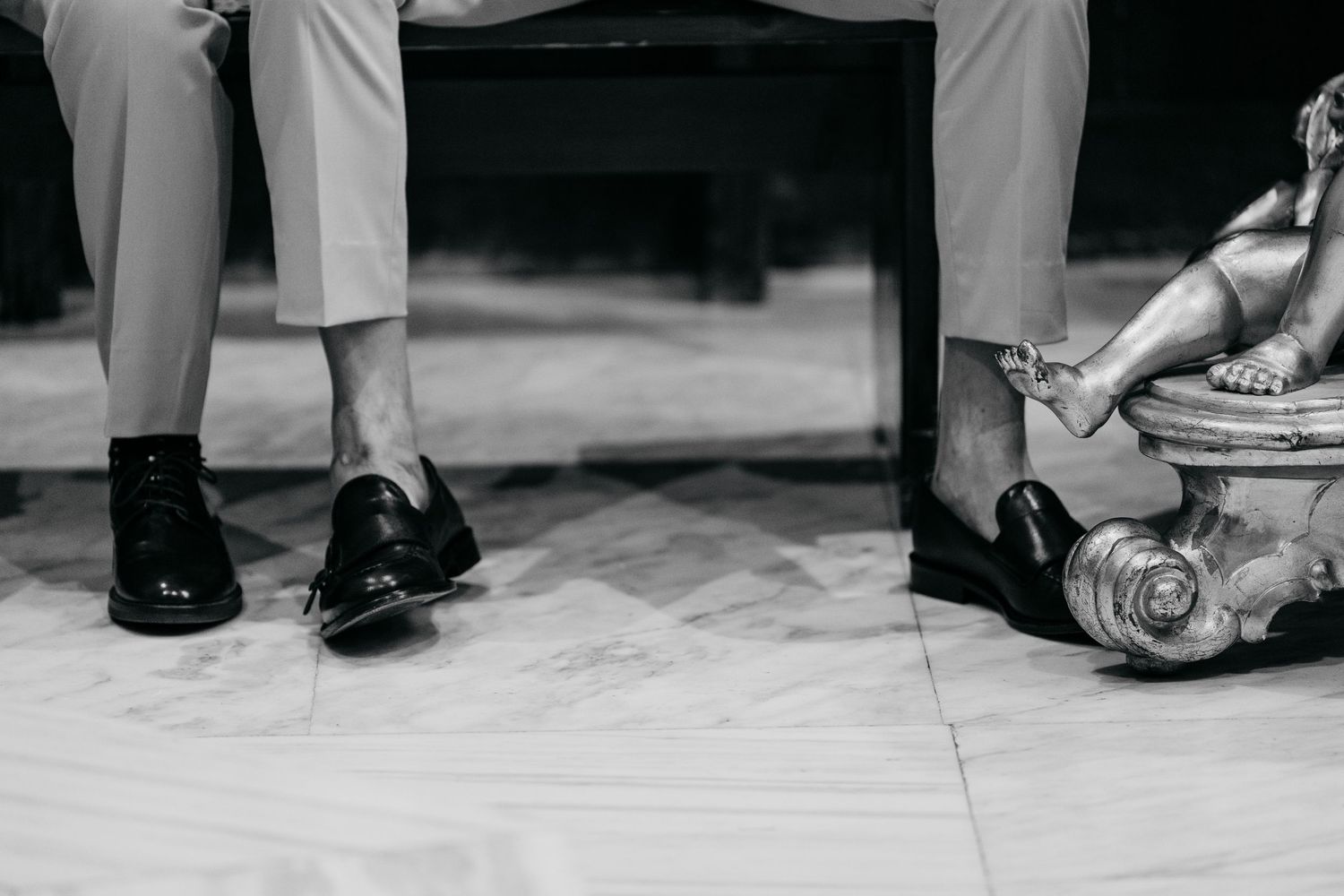 Black and white close-up of a groom's polished dress shoes and grey trousers seated on a chair at a wedding.