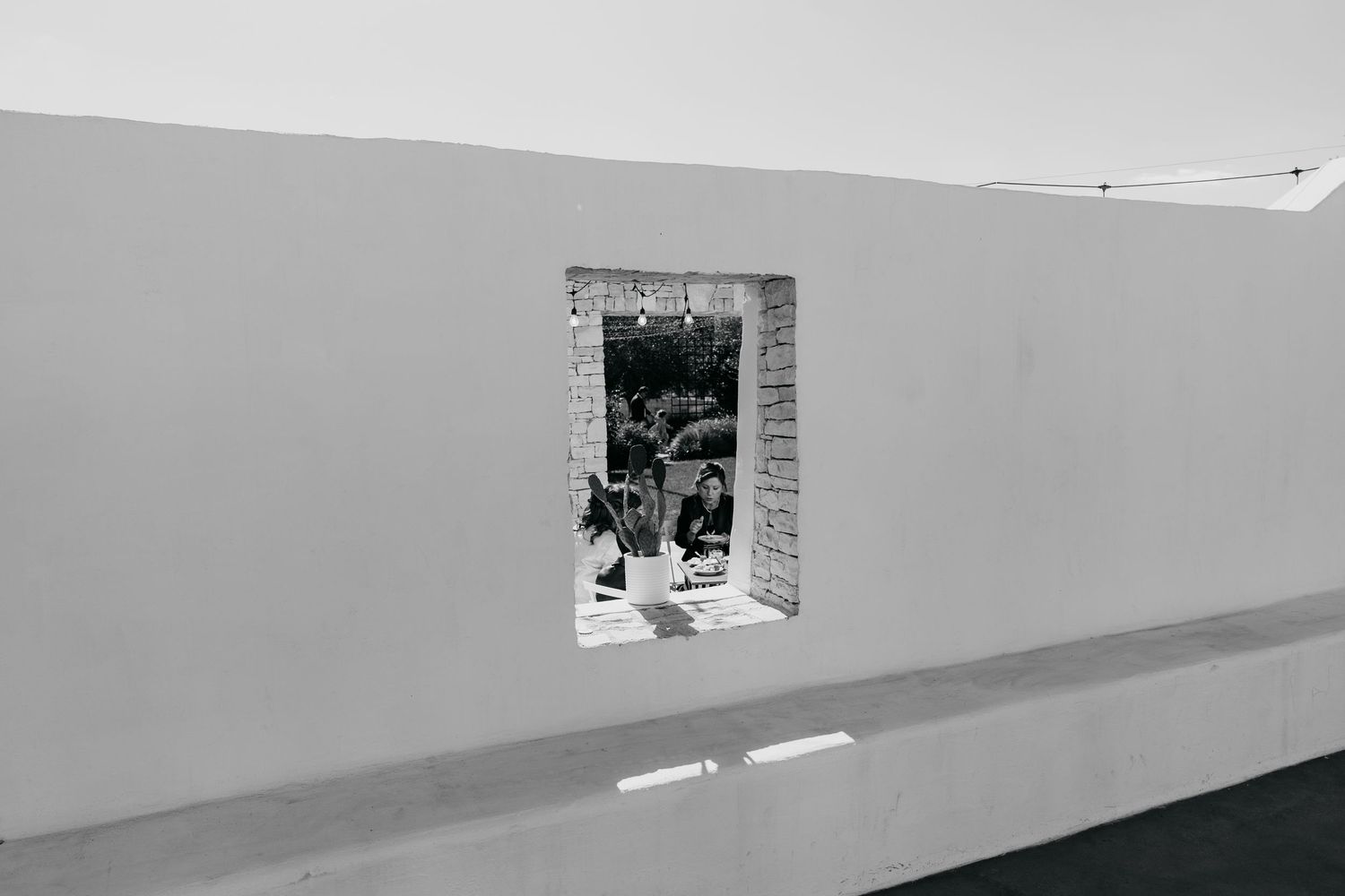 Black and white architectural photo of a white wall with a rectangular window revealing a garden scene beyond.