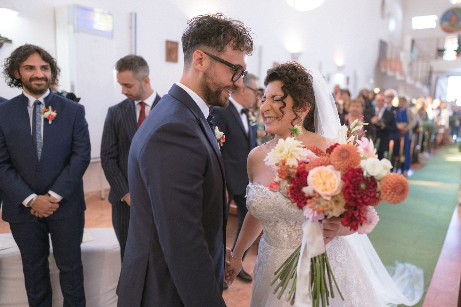 Groom in navy suit smiles at bride holding colorful bouquet during wedding ceremony in church aisle.