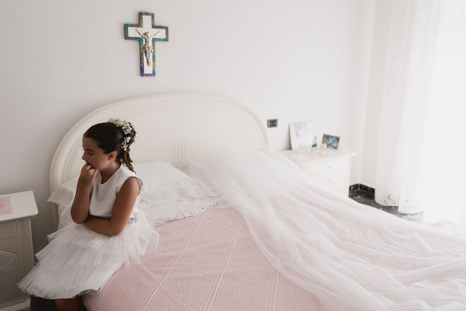 White wedding dress flowing across a bed with a cross hanging on the wall in a minimalist room.