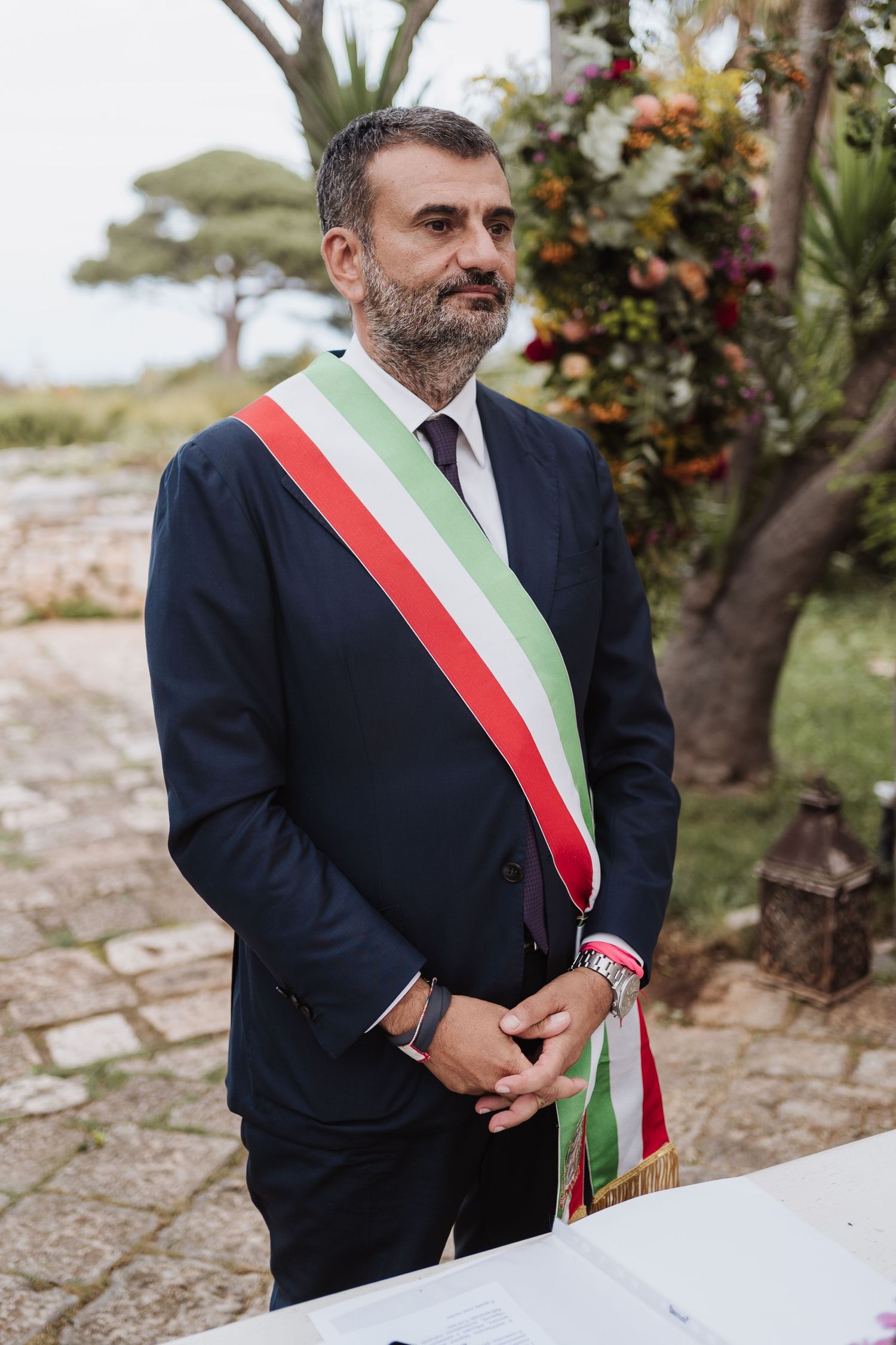 A government official wearing a dark suit with Italian tricolor sash stands in an outdoor setting.