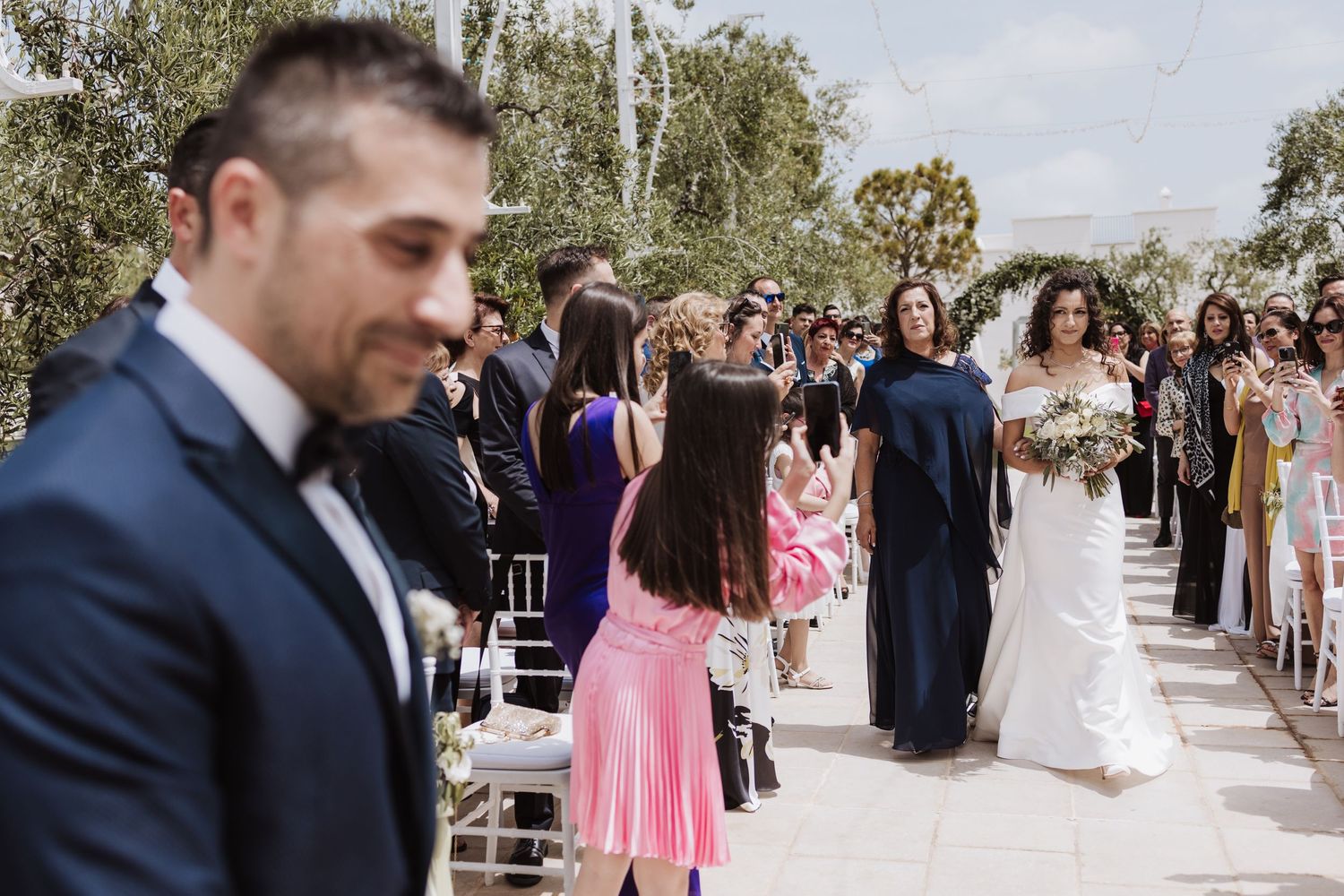 Outdoor wedding ceremony with guests seated on white chairs along a decorated aisle.