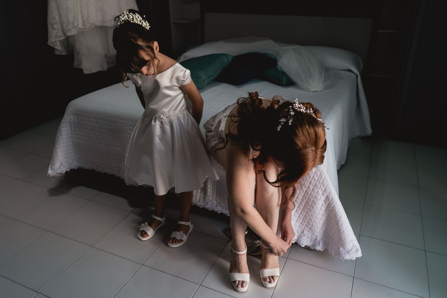 Two people in white dresses adjusting their shoes while sitting on a bed.