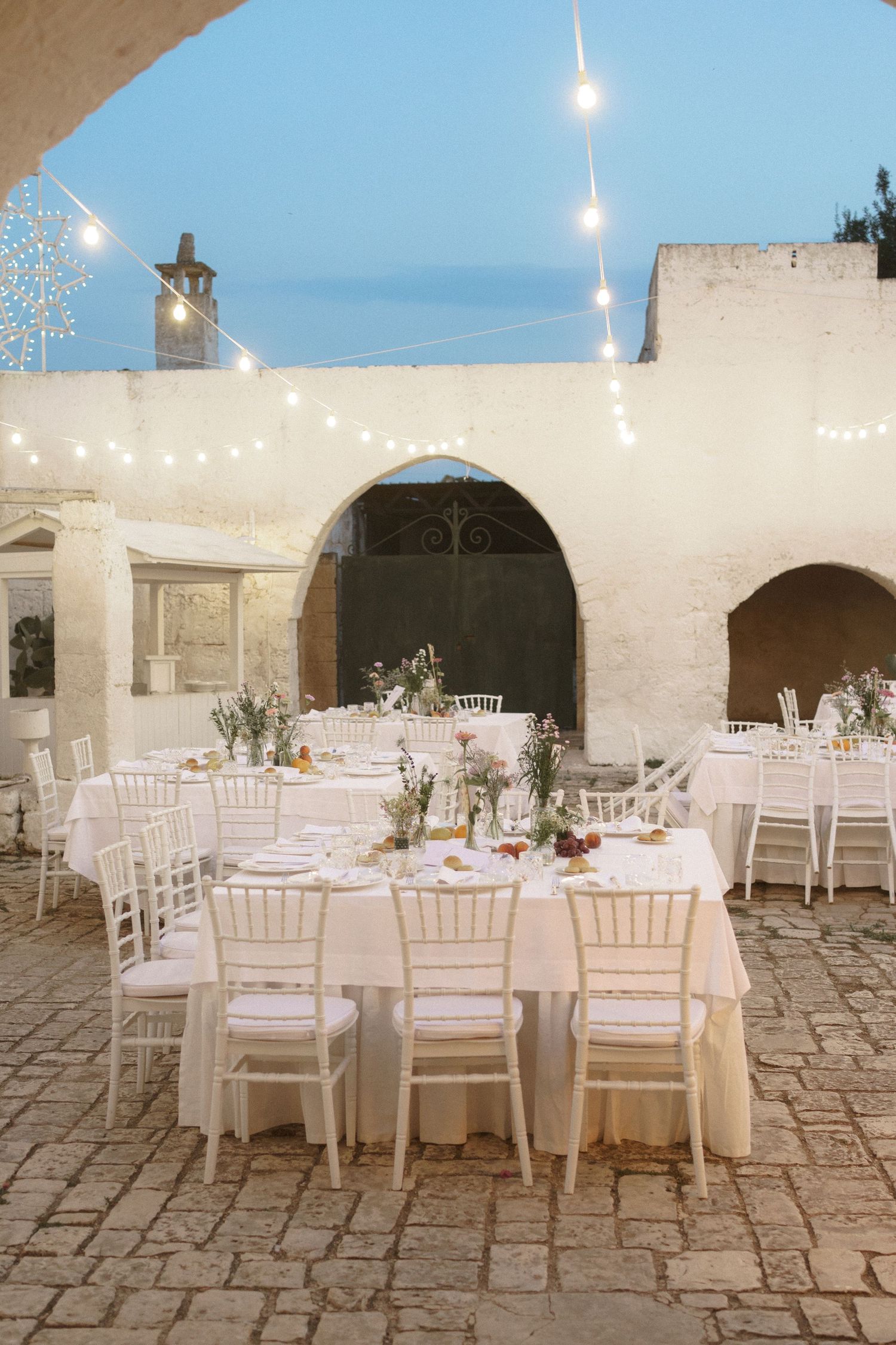 Elegant outdoor wedding reception setup with white tables and chairs under string lights at a Mediterranean-style venue.