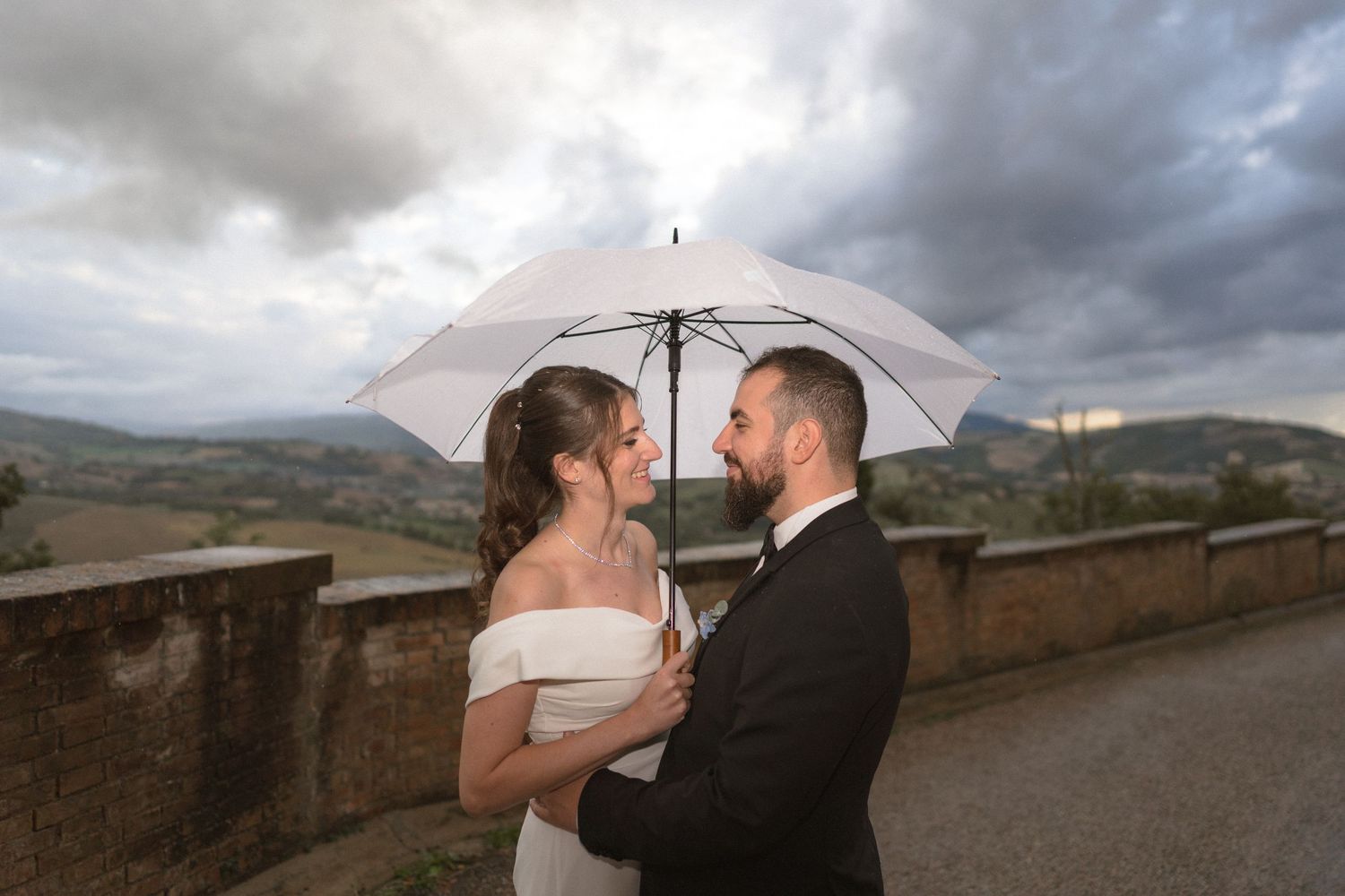 A romantic wedding couple shares an intimate moment under an umbrella against a dramatic cloudy landscape.