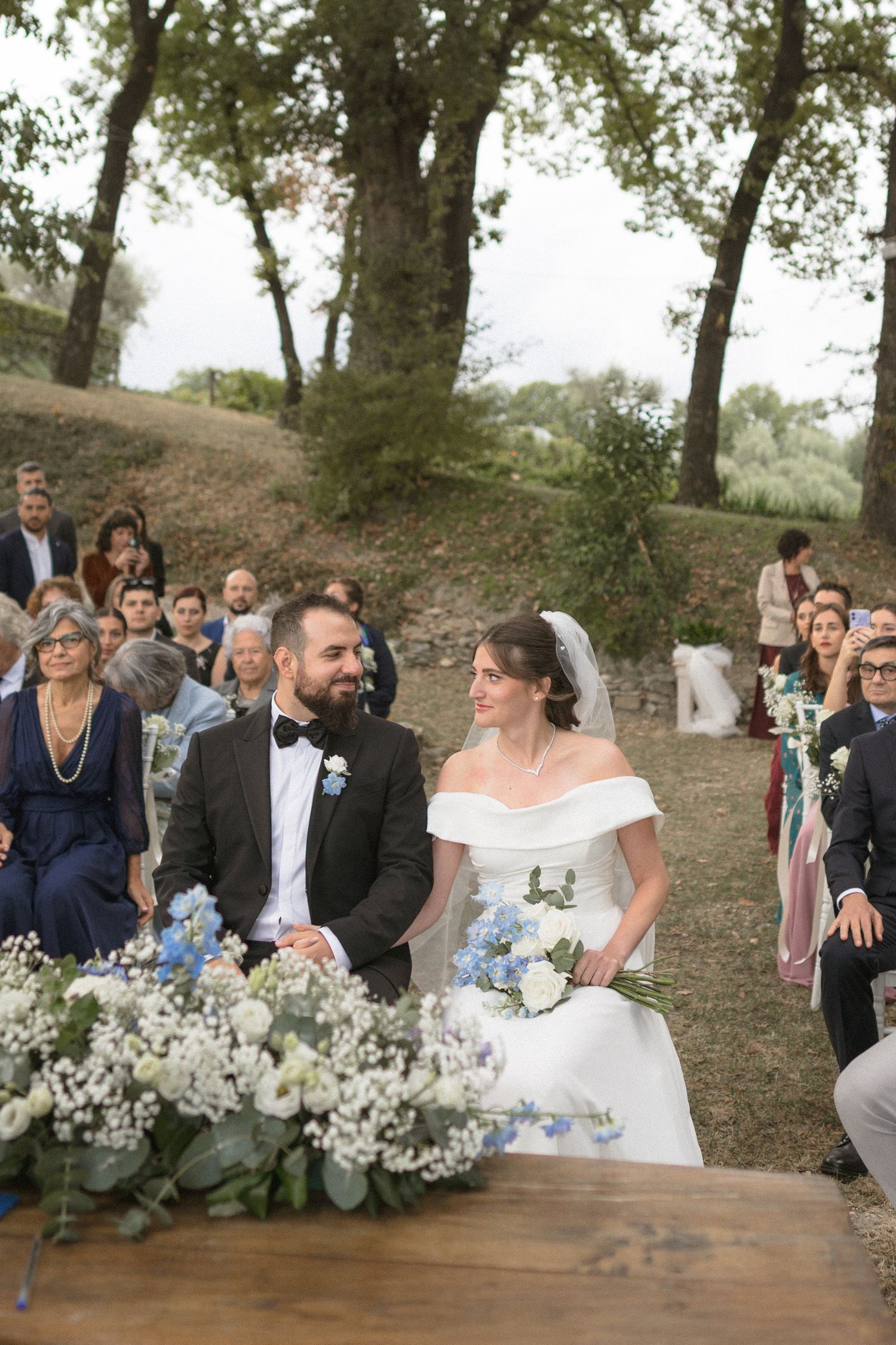 Outdoor wedding ceremony setup with white chairs and floral arrangements under trees.