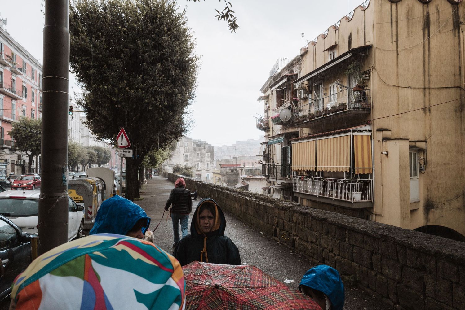 A rainy day street scene in Naples with people walking under umbrellas and stone buildings in the background.