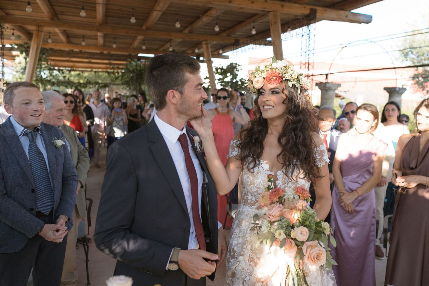 Outdoor wedding ceremony under a wooden pergola with floral decorations and gathered guests.