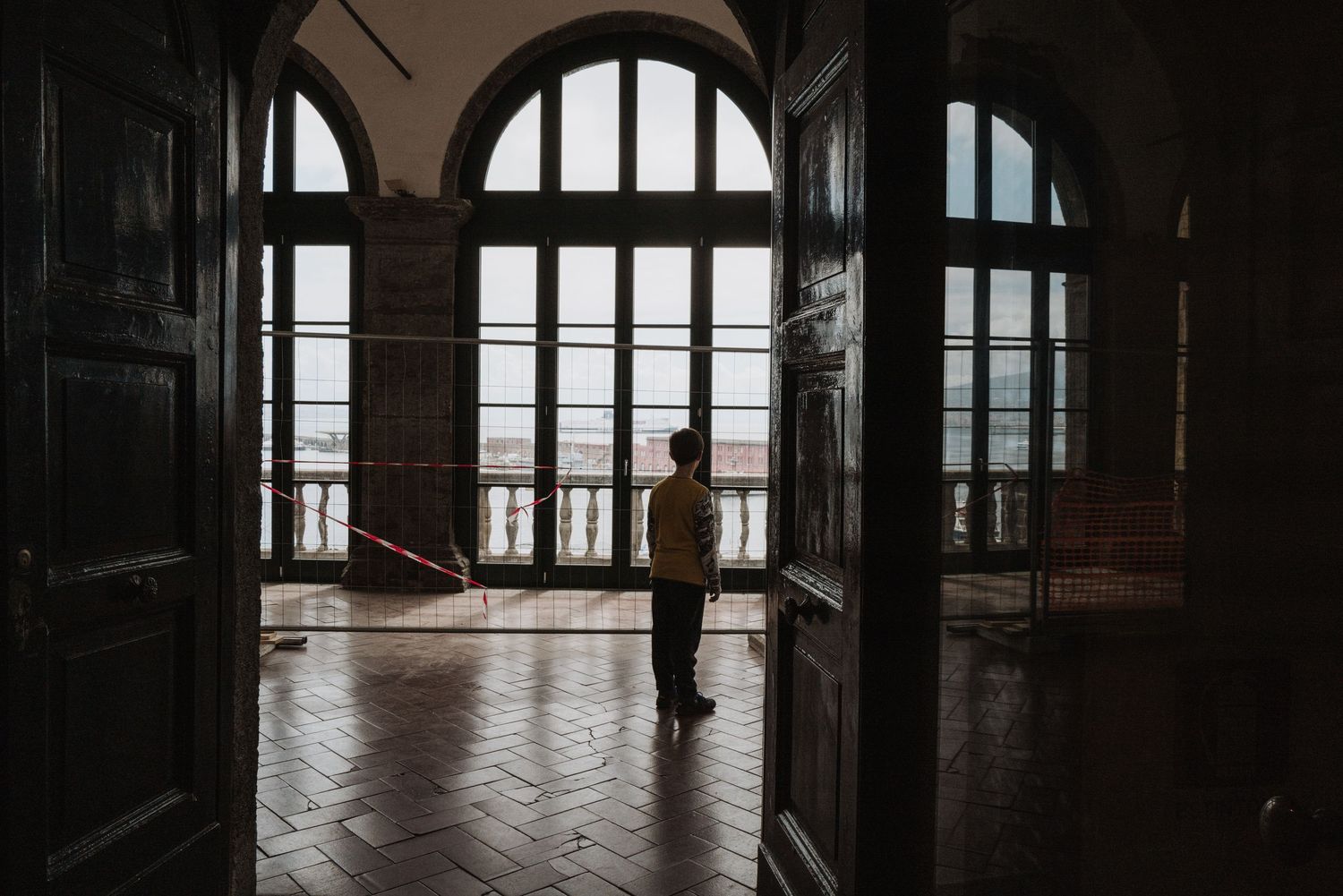 Silhouetted figure stands in arched doorway overlooking scenic view through grand windows in historic building.