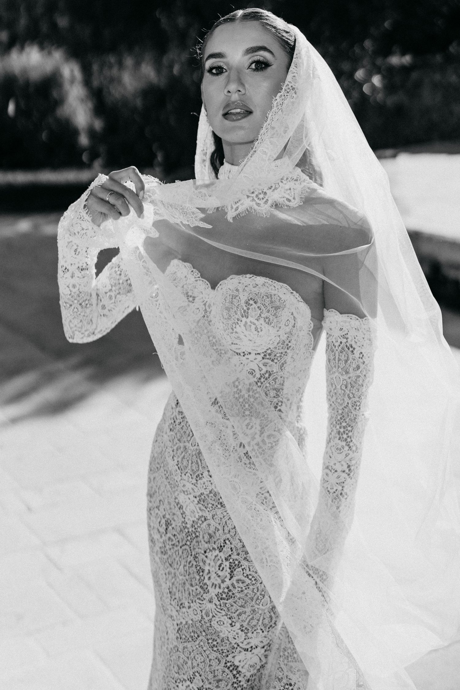 Black and white portrait of a bride in a long lace veil and sleeved lace gown lifting the sheer fabric.