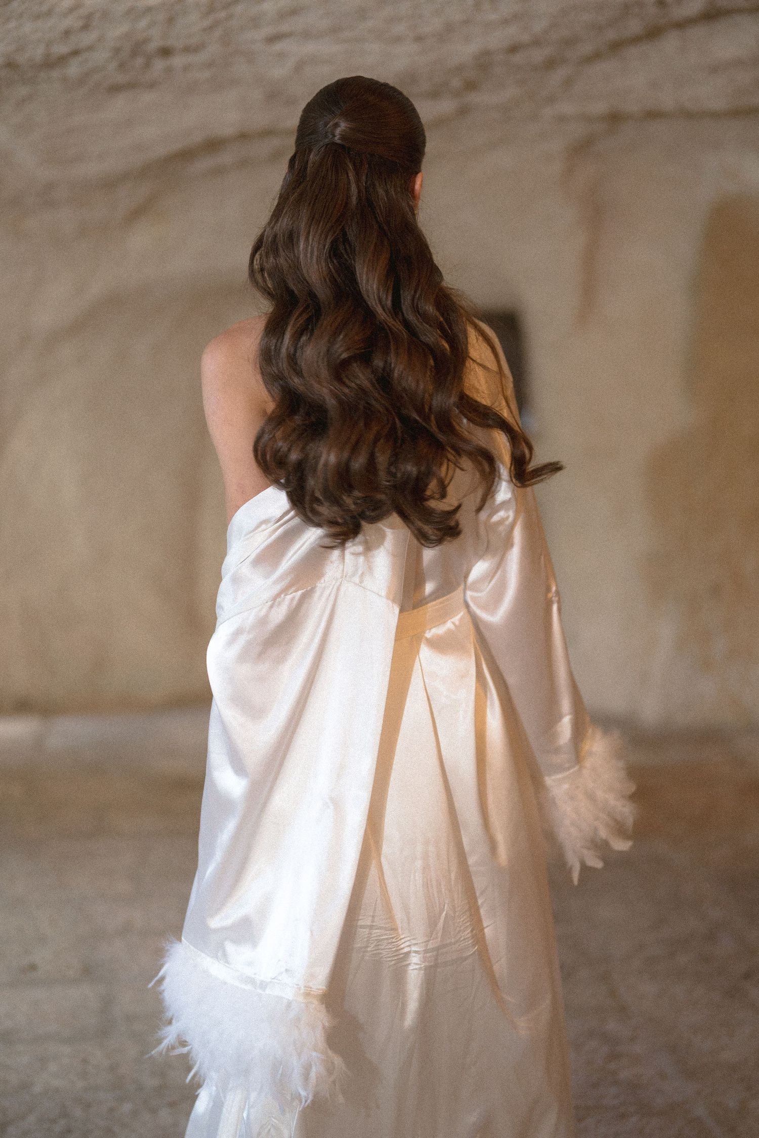 Bride seen from behind in a white satin robe with feather cuffs, long wavy brown hair styled in a half-up ponytail in a stone room.