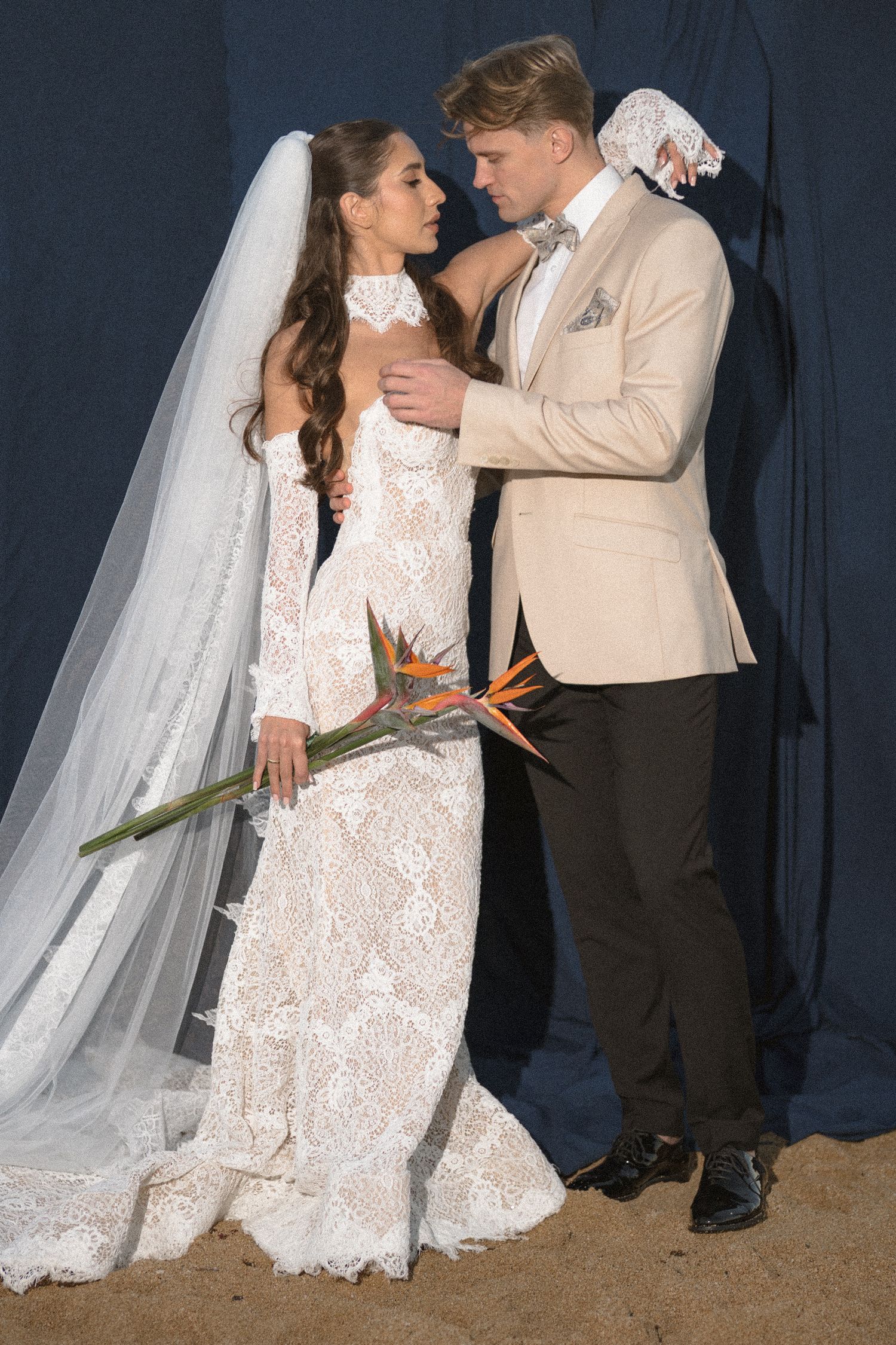 Bride in a fitted lace wedding dress and veil holding a bird of paradise bouquet while embracing her groom in a cream blazer.