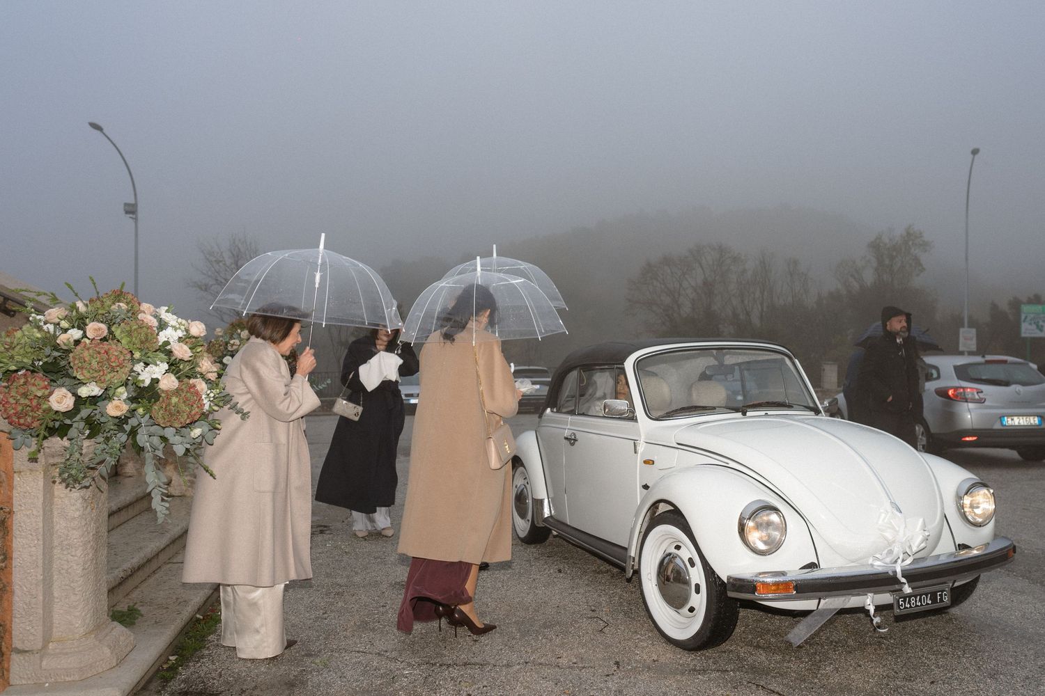 Guests with transparent umbrellas gather near a vintage white VW Beetle convertible on a foggy wedding day.