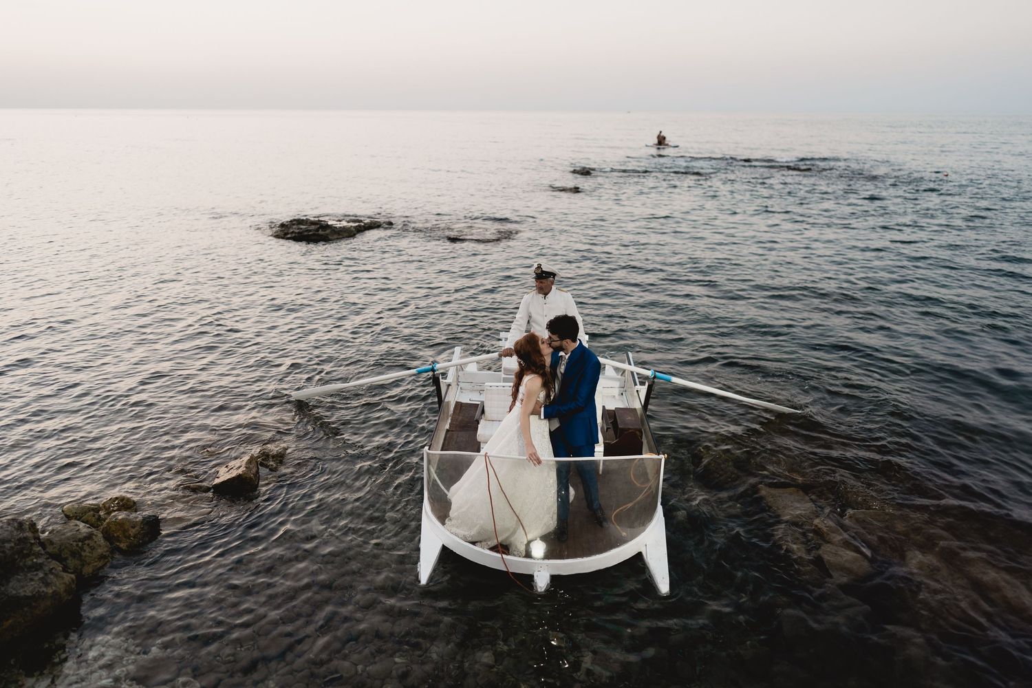 A couple stands at the bow of a small boat near a rocky coastline at dusk with a lighthouse in the distance.
