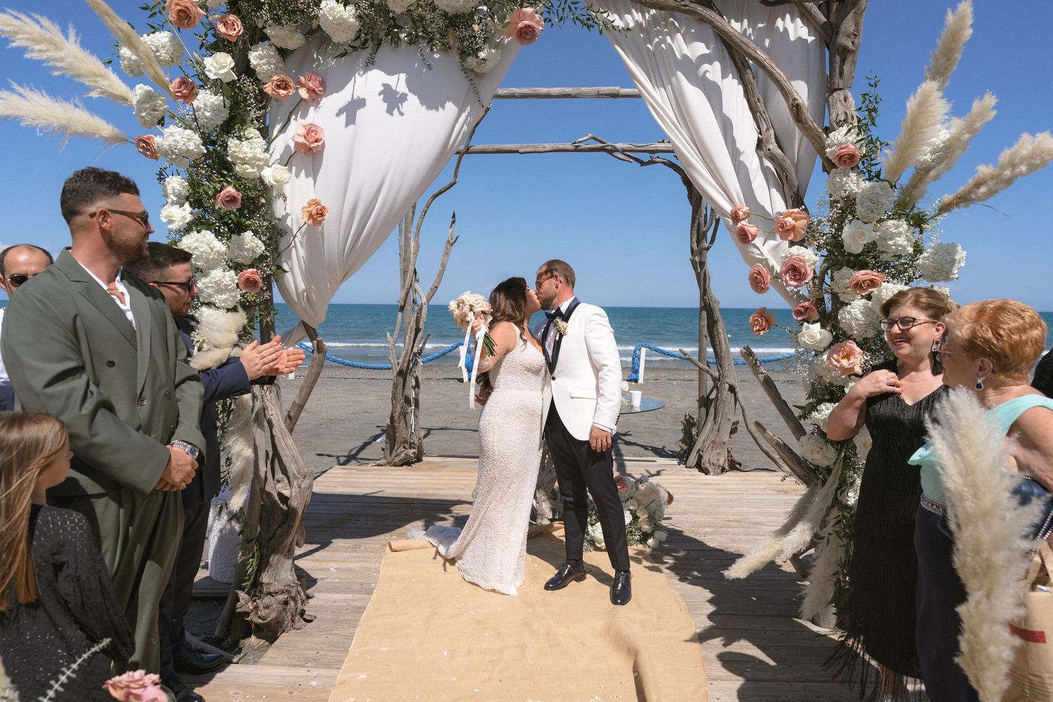 Beach wedding ceremony under a floral arch with white draping and ocean backdrop.