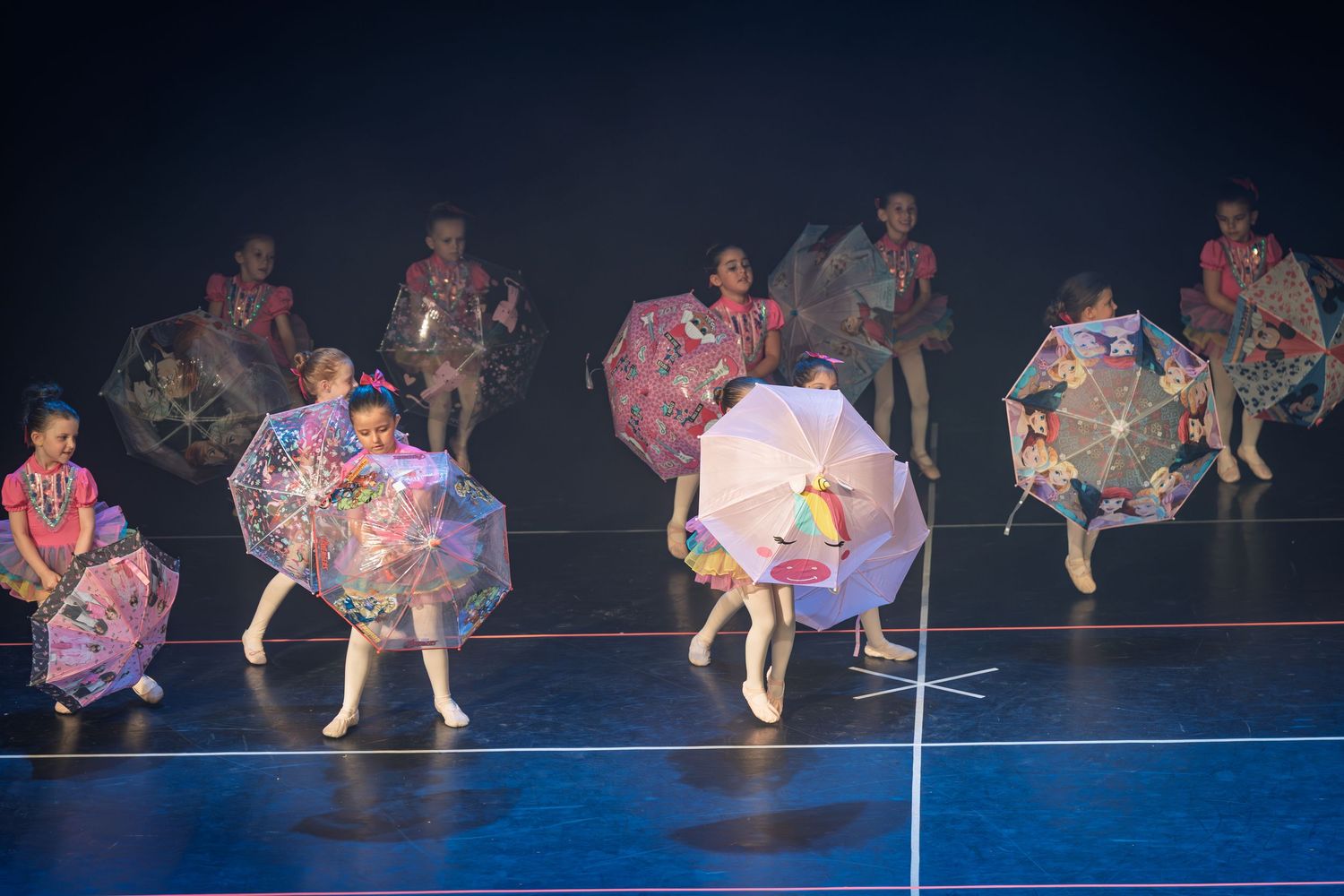 Dancers in colorful ruffled skirts perform a synchronized routine on a dark stage with dramatic lighting.