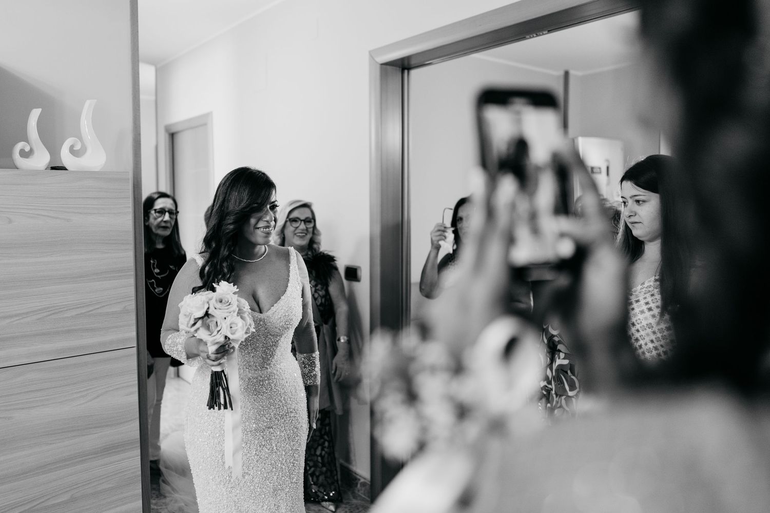 A candid black and white moment of a bride in a lace gown being photographed in a hallway.