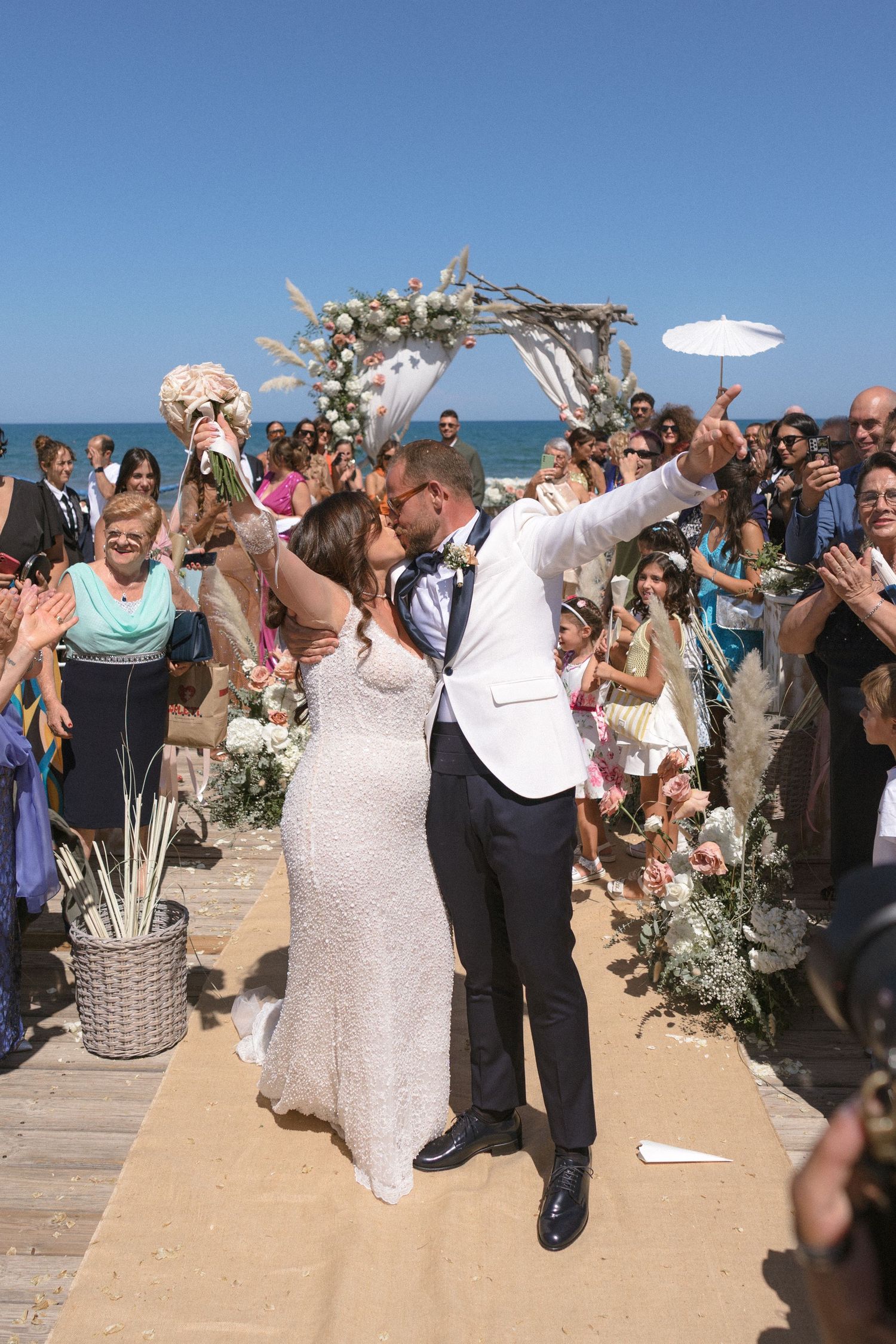 Newlyweds celebrate under a driftwood arch decorated with flowers at their beach wedding.