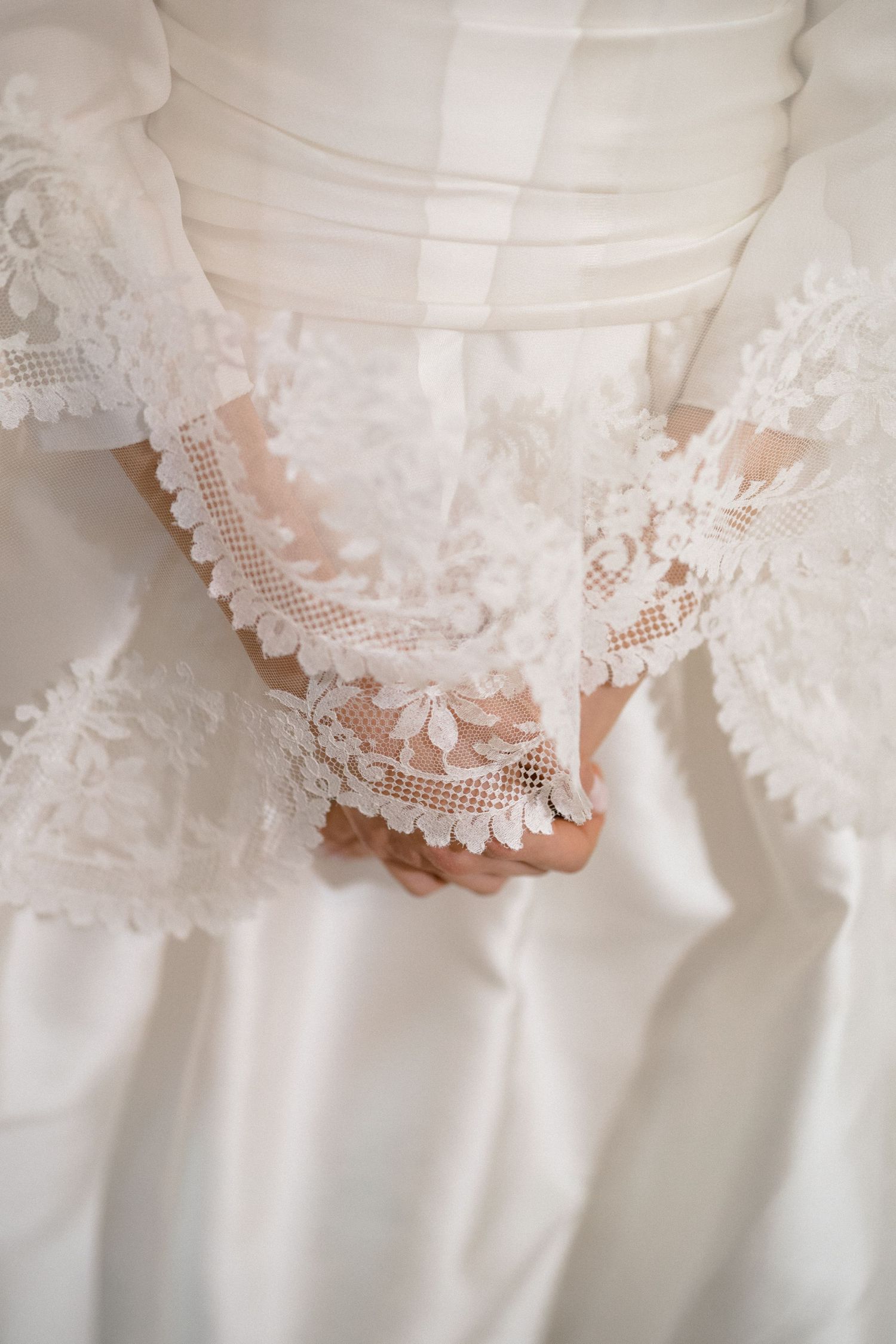 Close-up of a bride's lace-trimmed wedding dress sleeves with hands clasped together.