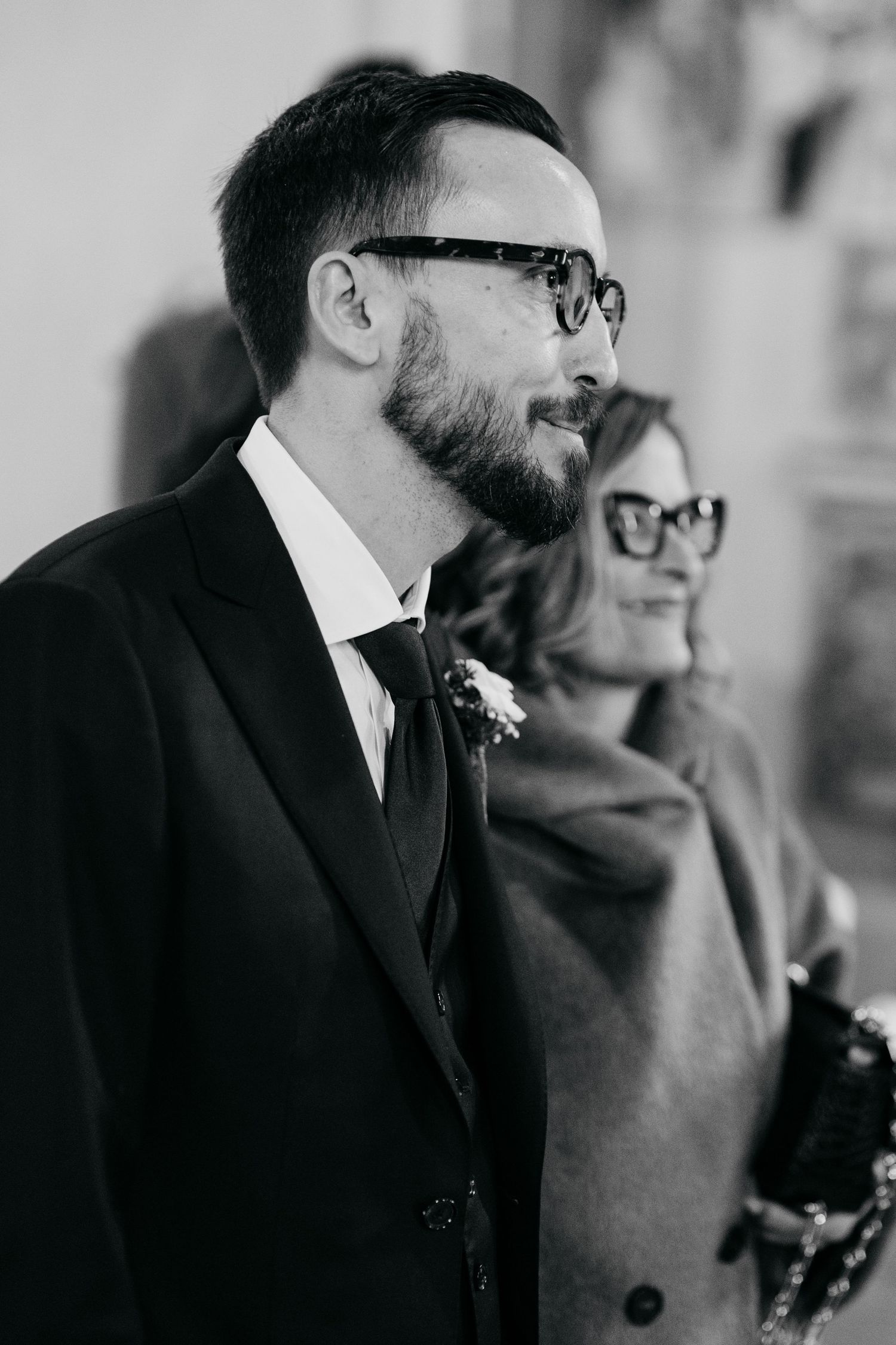 A bearded groom wearing glasses and a boutonniere stands at the altar in black and white.