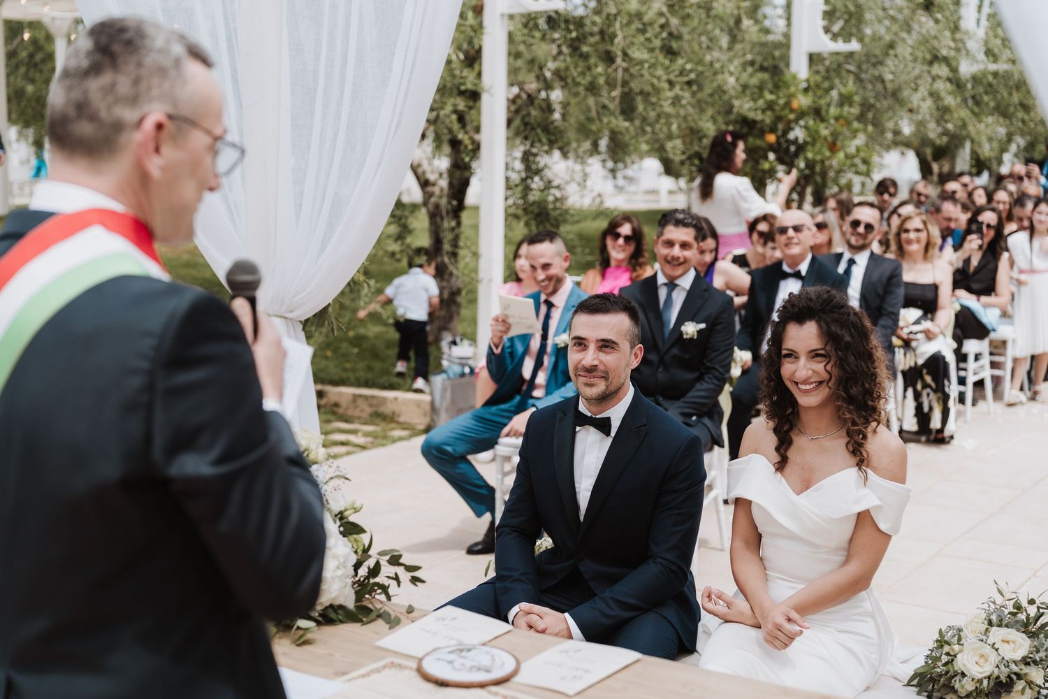 A wedding ceremony in progress with guests seated outdoors under an elegant white canopy.